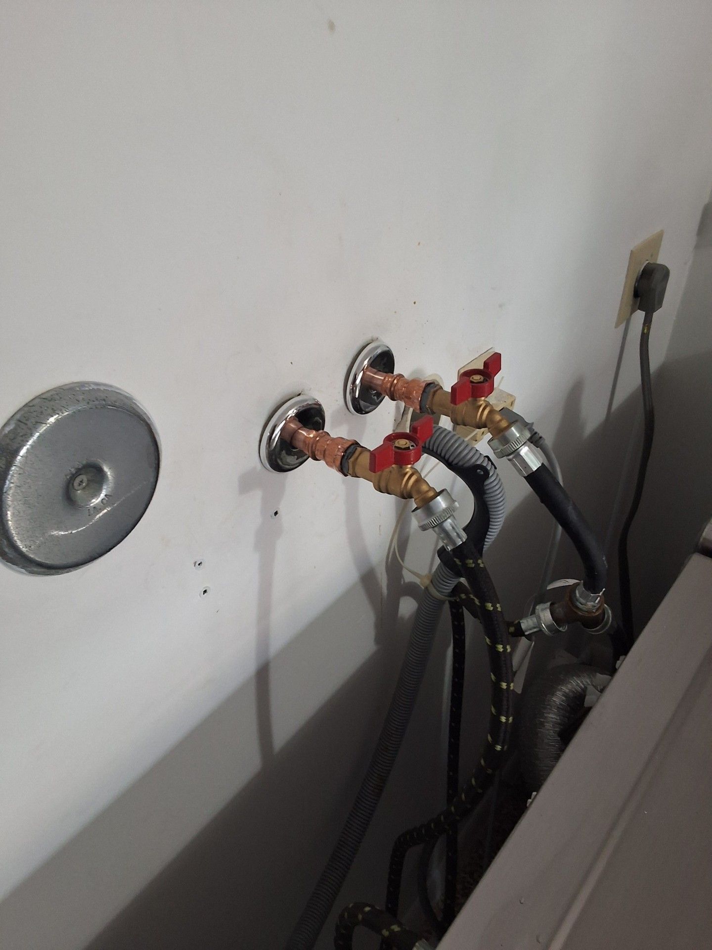Plumbing valves and hoses mounted on a white wall beside a utility fixture