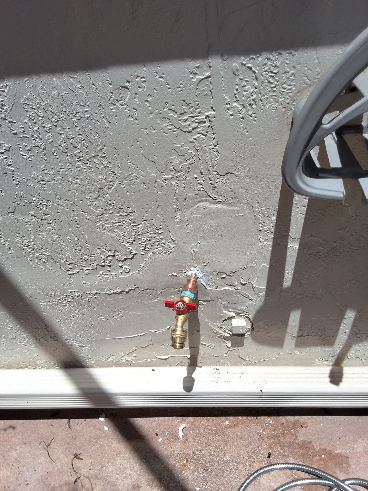 Outdoor wall with a small brass water spigot and red handle below a gray chair shadow.