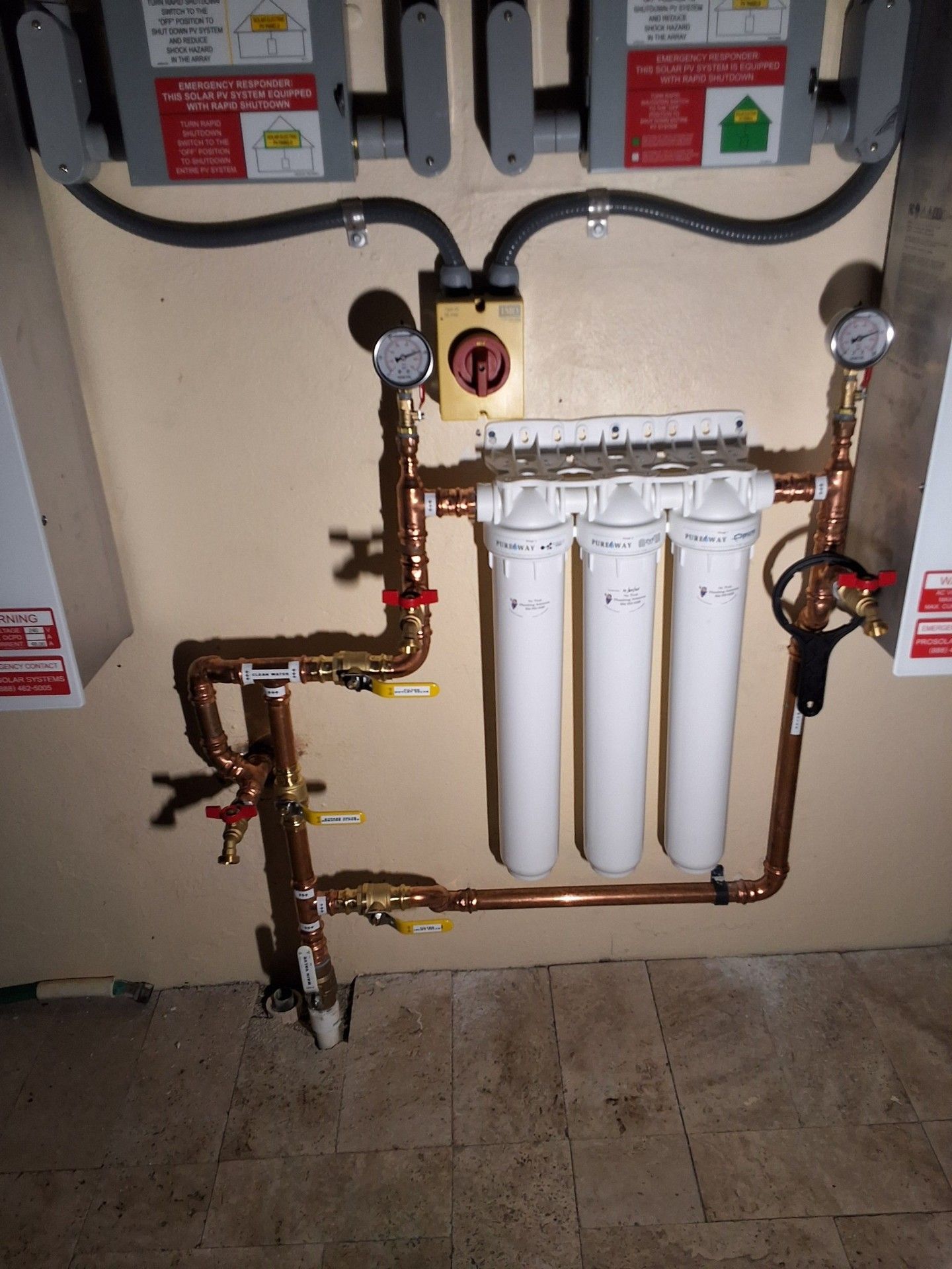 Wall-mounted water filtration system with white filter cartridges, copper pipes, and gauges in a utility room