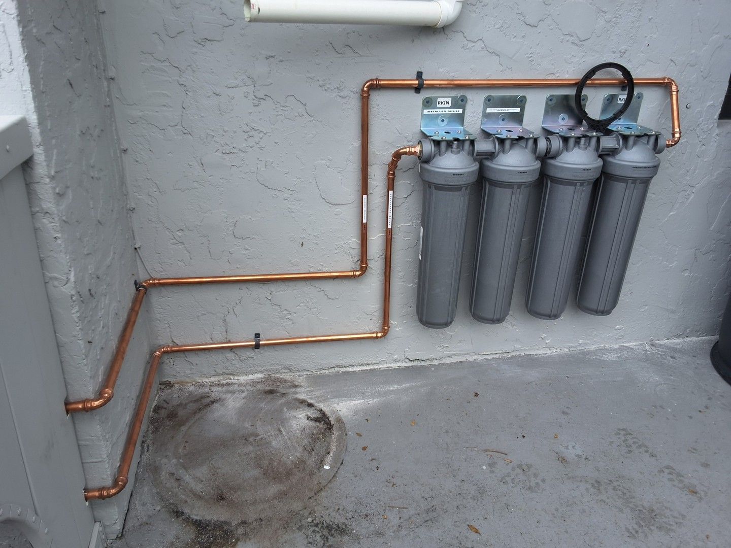 Copper pipes and four gray water filters mounted on a gray wall above a concrete floor.