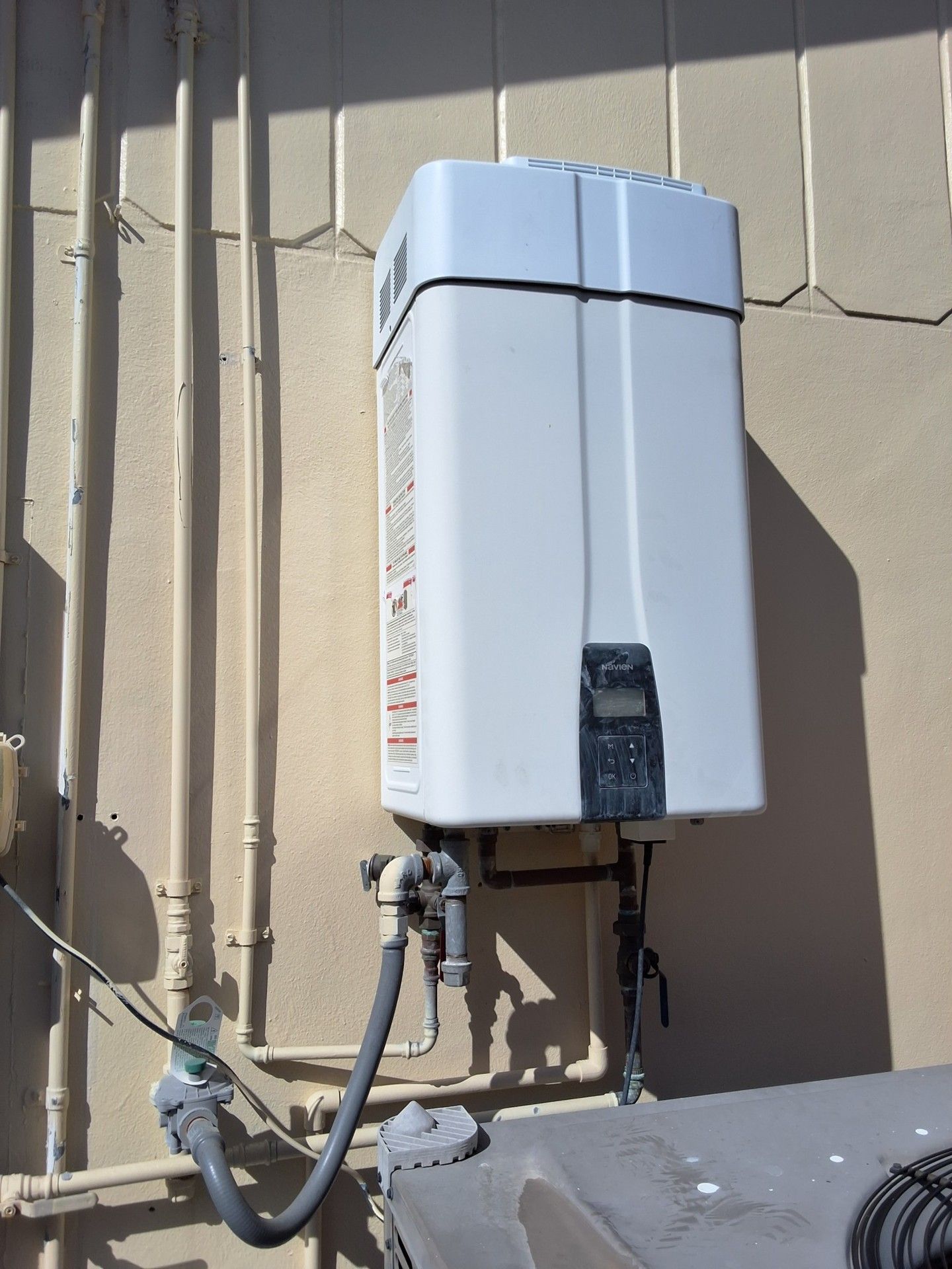 White wall-mounted tankless water heater on an exterior stucco wall with visible pipes and conduit