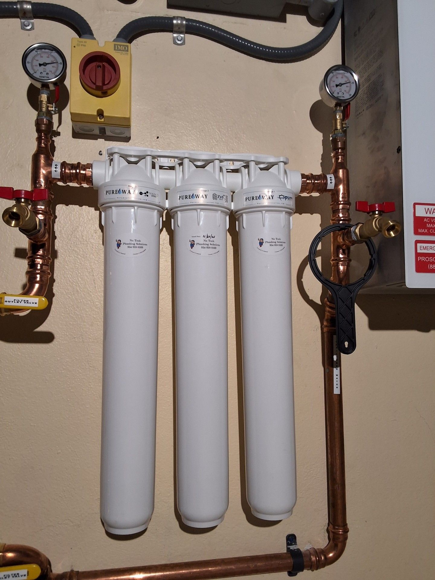 Three white water filters with gauges and copper pipes on a utility wall