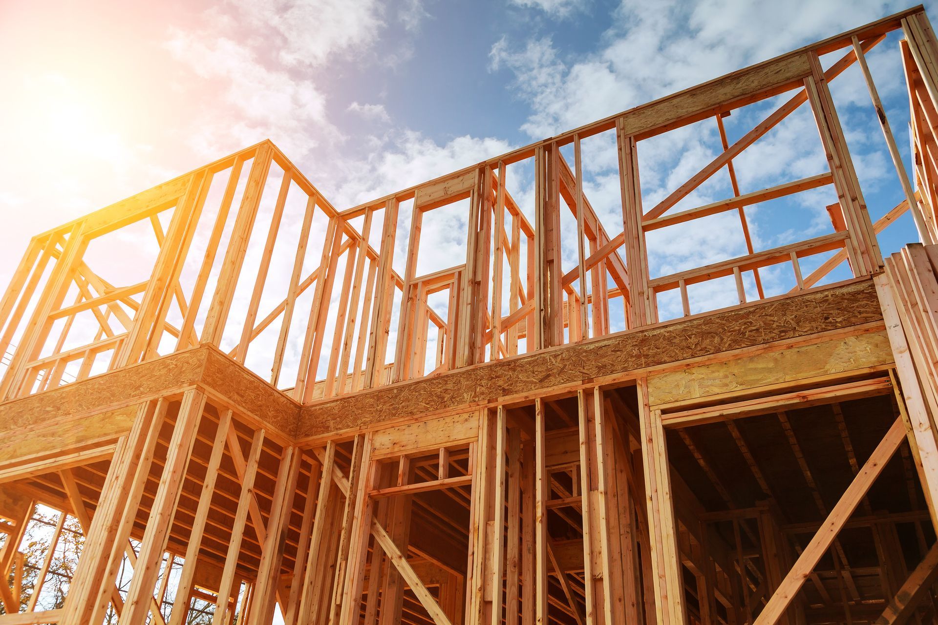 Wooden house framing under construction against a bright blue sky with sun glare