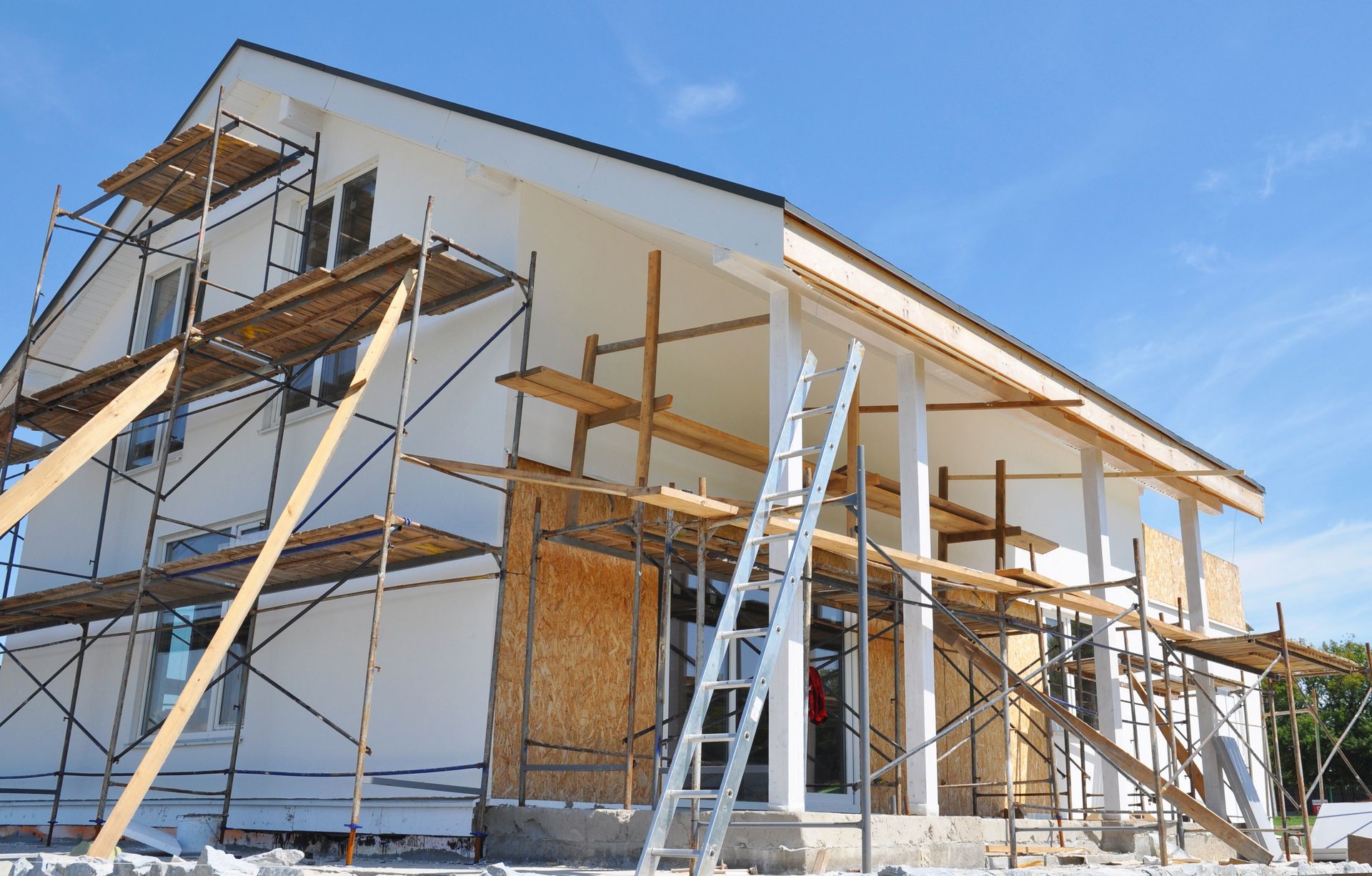 House under construction with wooden scaffolding, ladders, and white siding under a clear blue sky