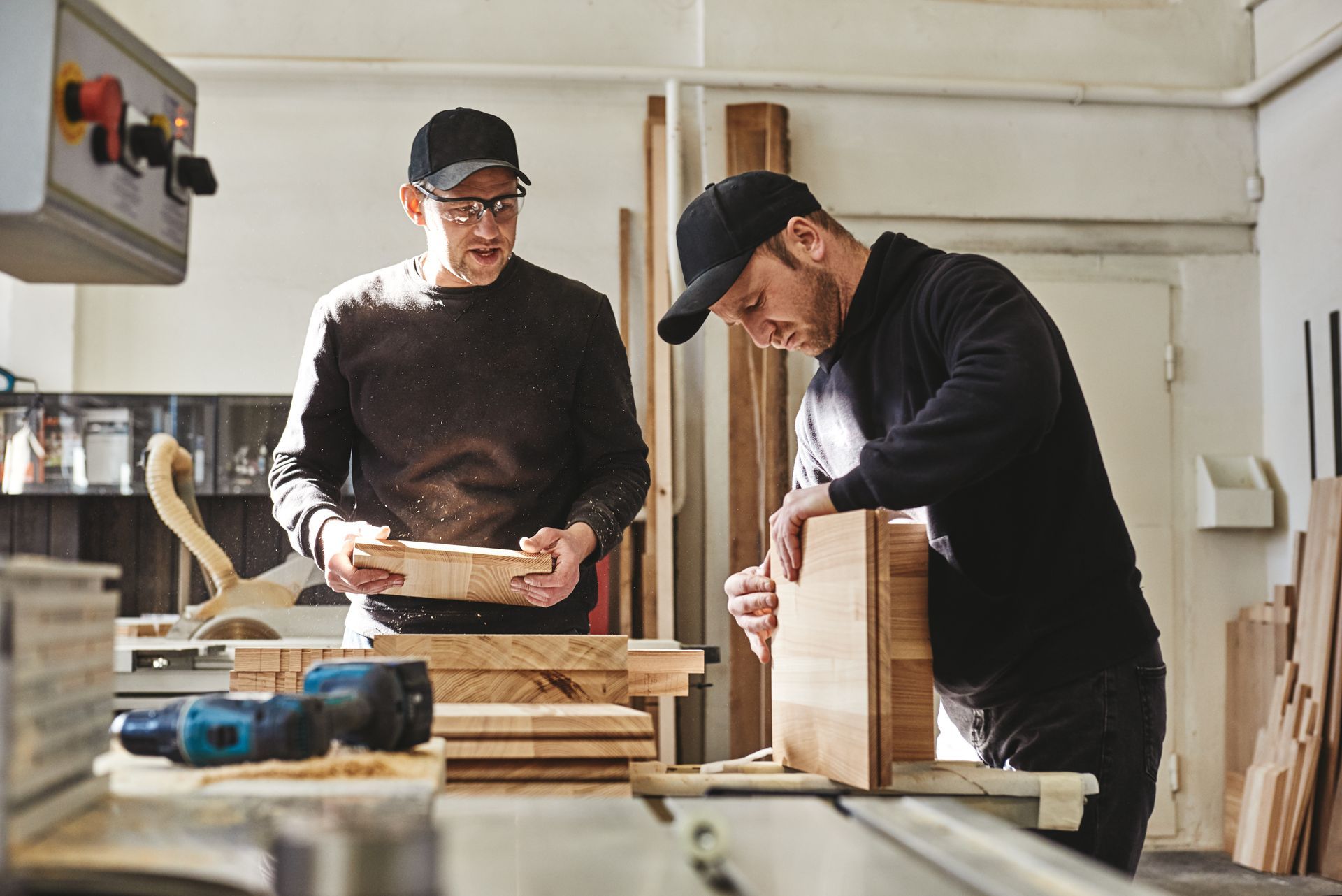Two people woodworking at a workbench in a bright workshop, measuring and assembling wood pieces.