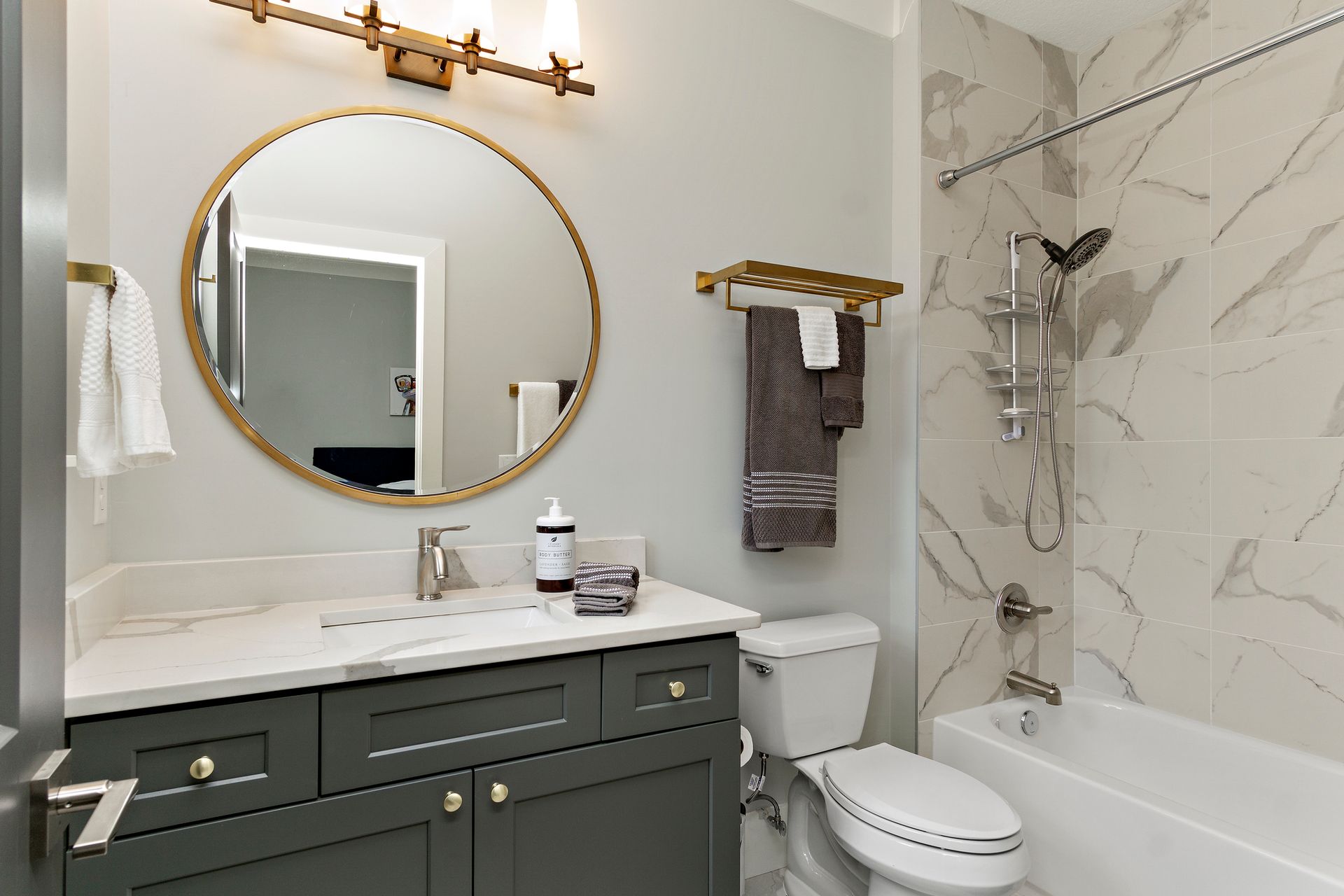 Bright modern bathroom with gold fixtures, round mirror, dark vanity, and white tub shower.