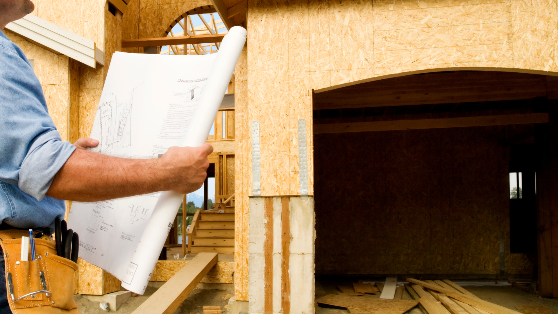 A man is holding a blueprint in front of a house under construction.