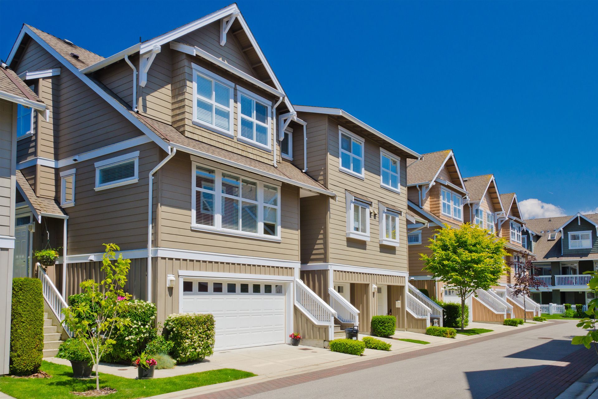 Row of beige townhouses with white trim and garages on a sunny street.