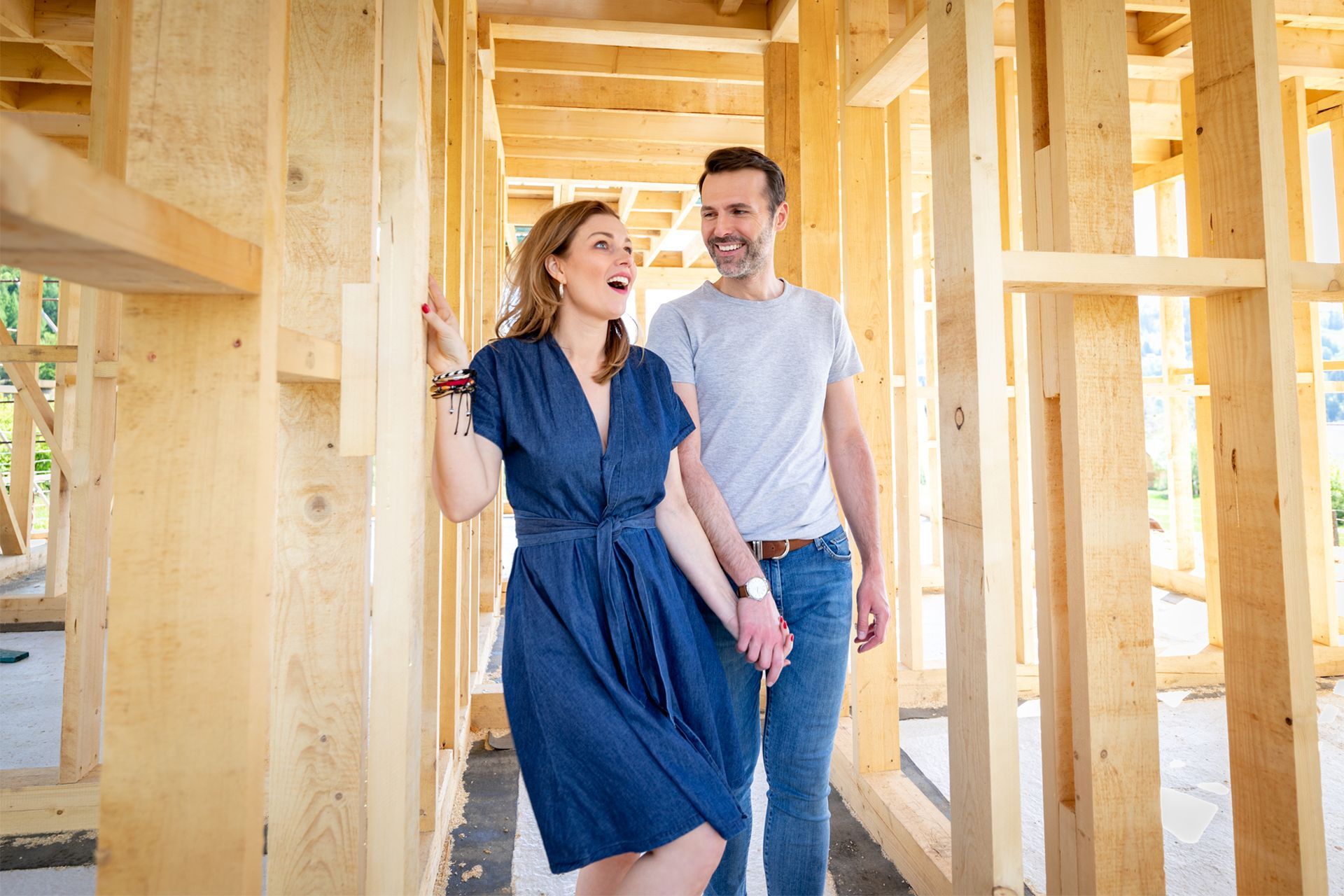 Couple holding hands, looking excitedly at the wooden frame of a new house under construction.