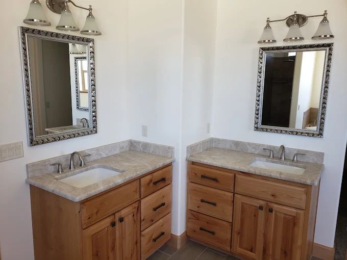 Two bathroom vanities with mirrors, lighting, sinks, and wooden cabinets against white walls.