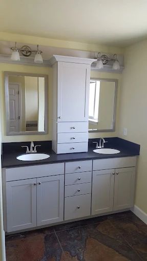 Bathroom with double sinks, white cabinets, black countertop, tall storage, mirrors, and light fixtures.