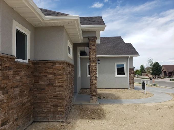 A house exterior with stone accent, grey stucco, white trim, and dark roof under a blue sky.