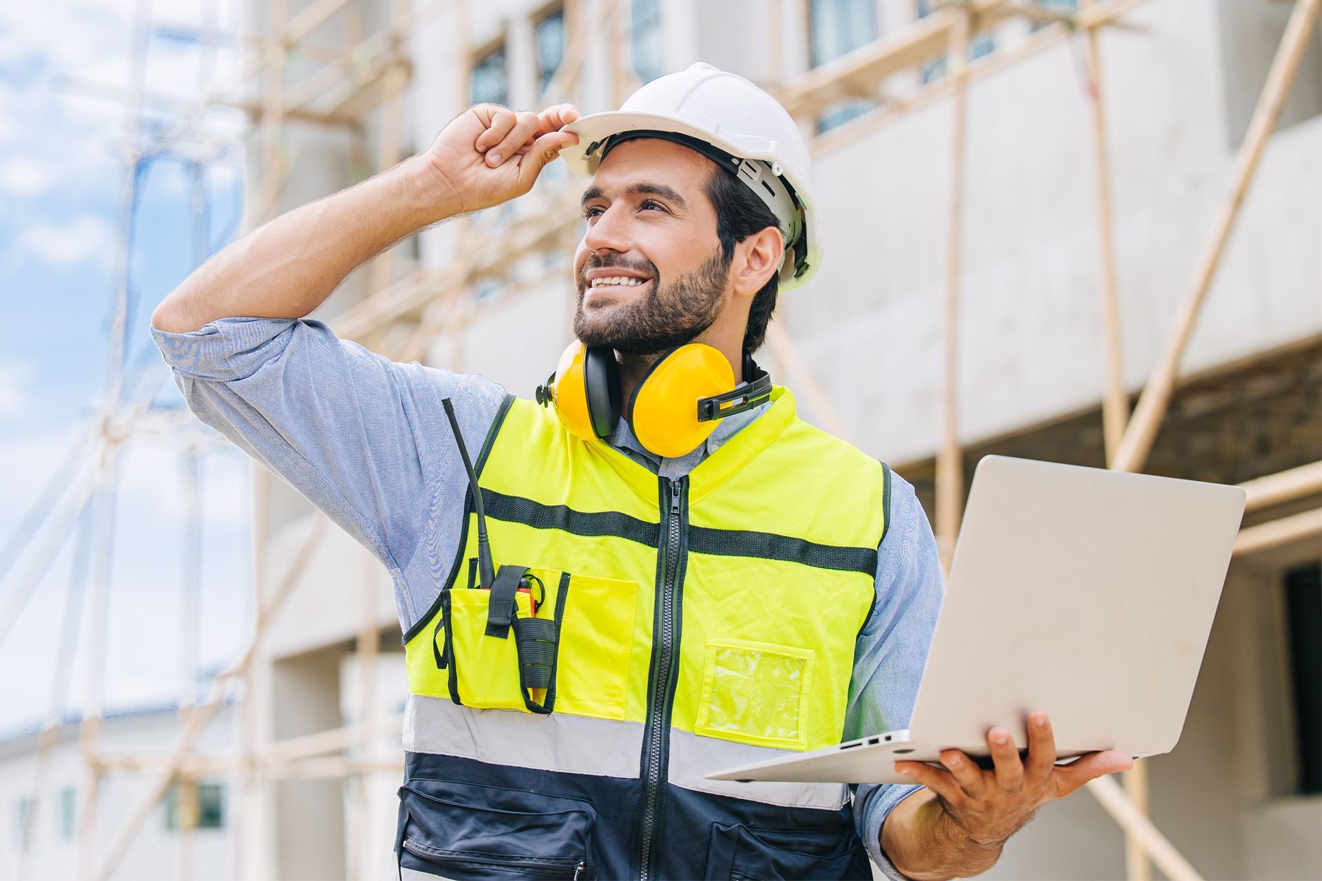 Construction worker in hard hat and vest, smiling, holding laptop at a construction site.