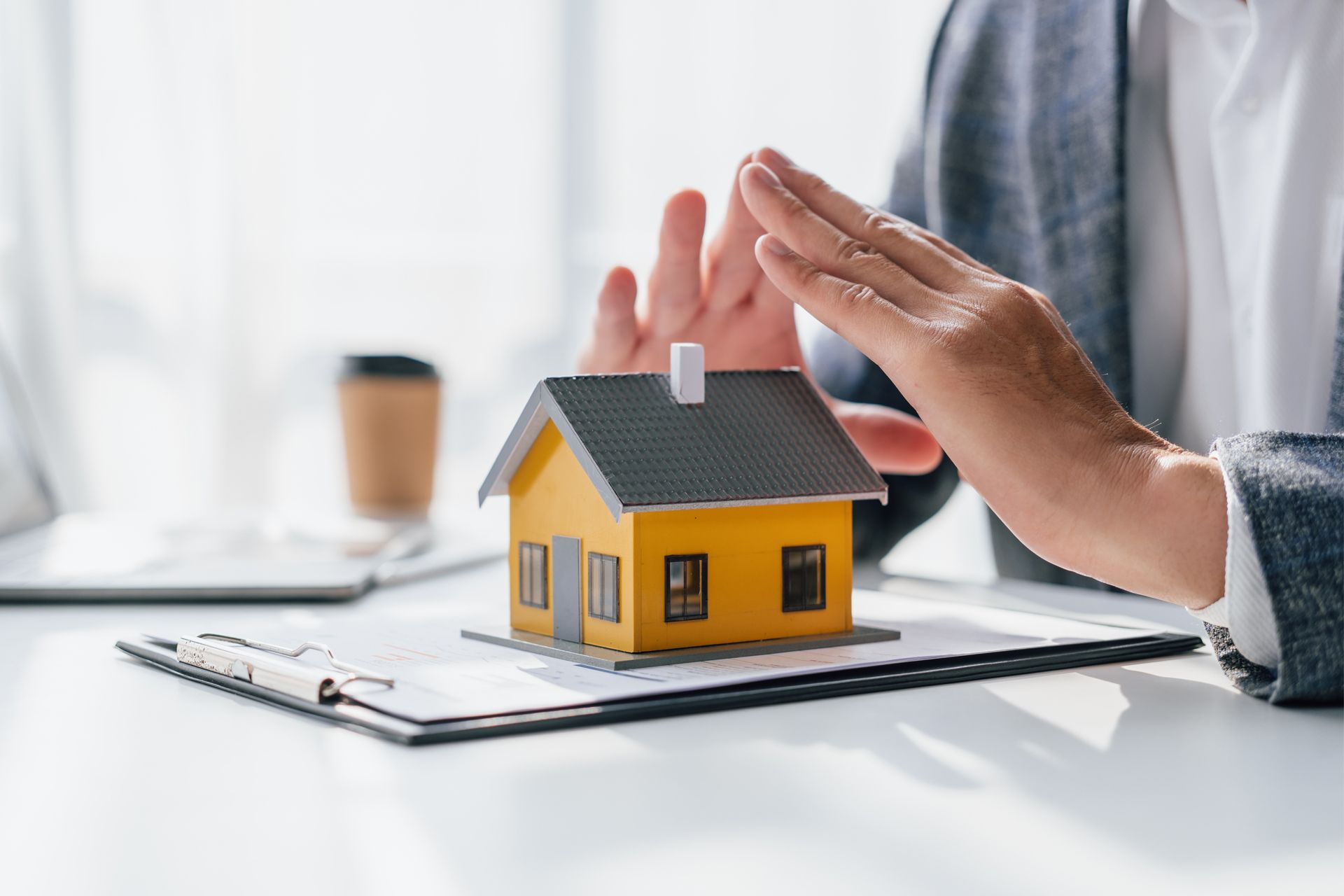 Person's hands protecting a miniature yellow house on top of a clipboard with paperwork and a coffee cup.