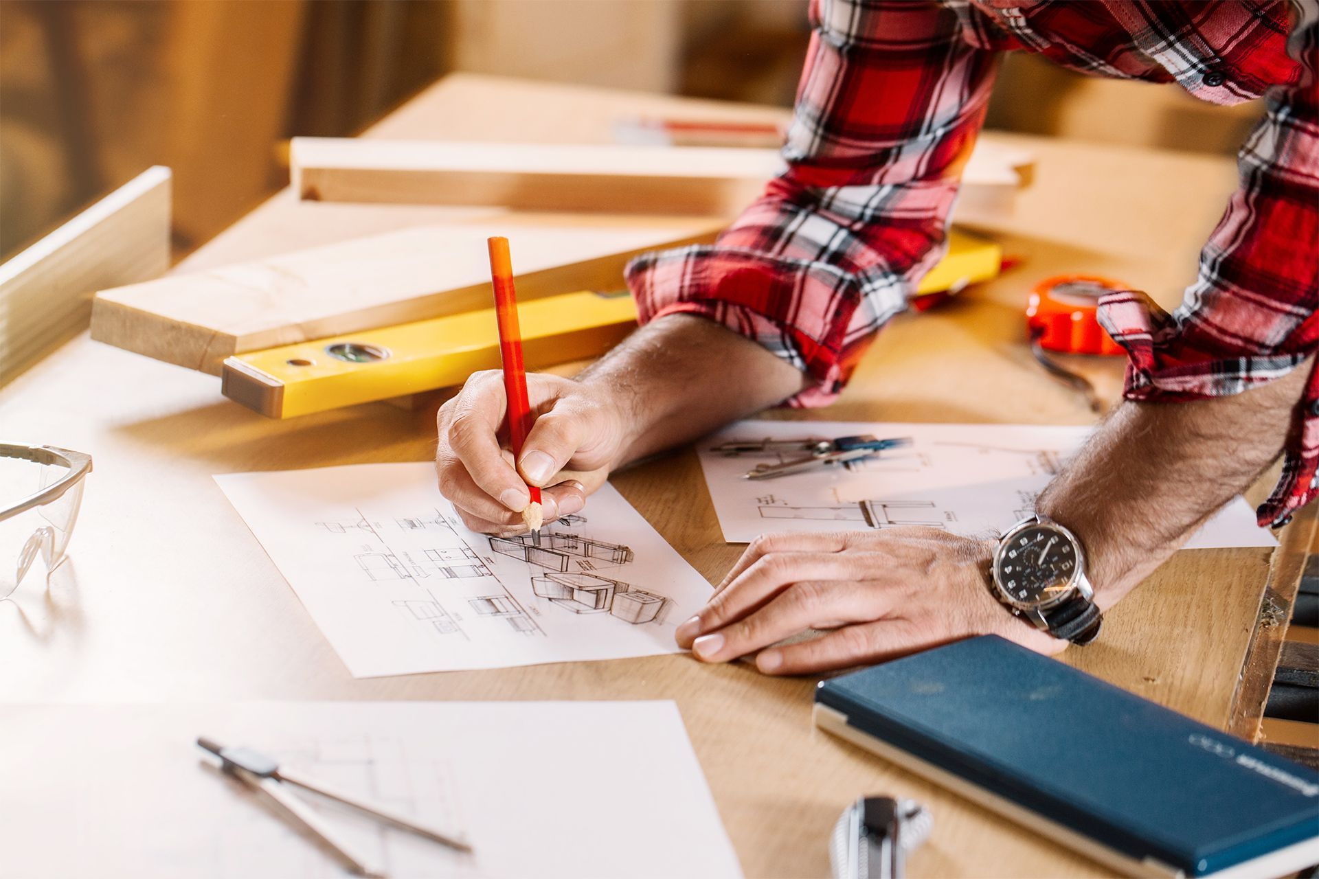 Person in plaid shirt sketching blueprints at a workbench, with tools like a level, tape measure, and compass.
