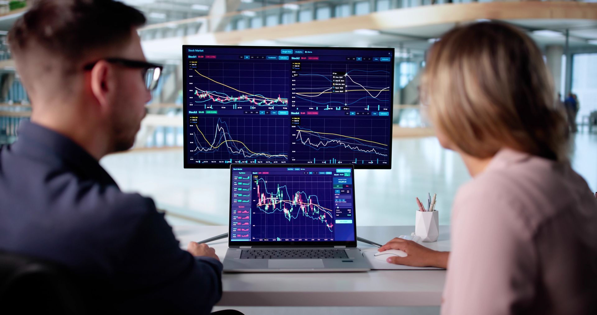 Two people analyzing financial charts on dual monitors and a laptop in a modern office setting