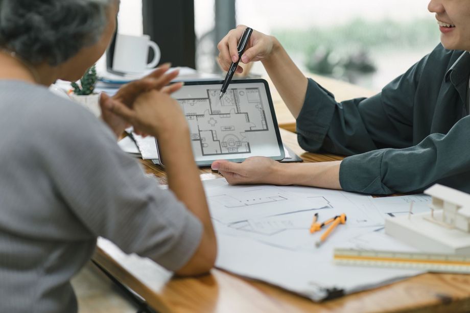 Person points at floor plan on tablet, discussing it with another person at a table.