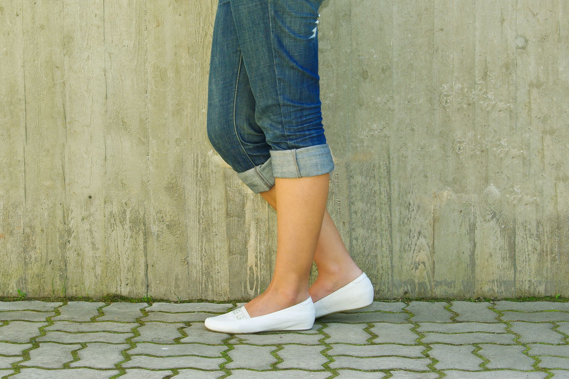 Person wearing cuffed blue jeans and white flats stands on a brick surface in front of a concrete wall.