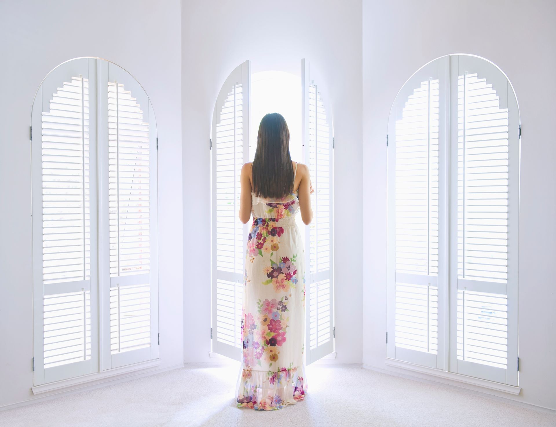 Woman in floral dress standing by arched white shutters, one open to light.