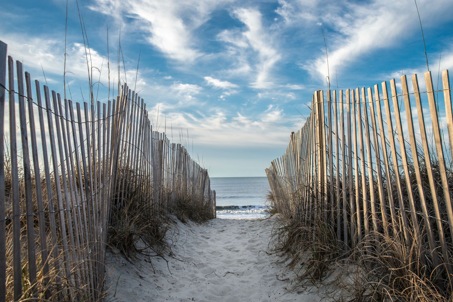 A wooden fence along a sandy path leading to the ocean.