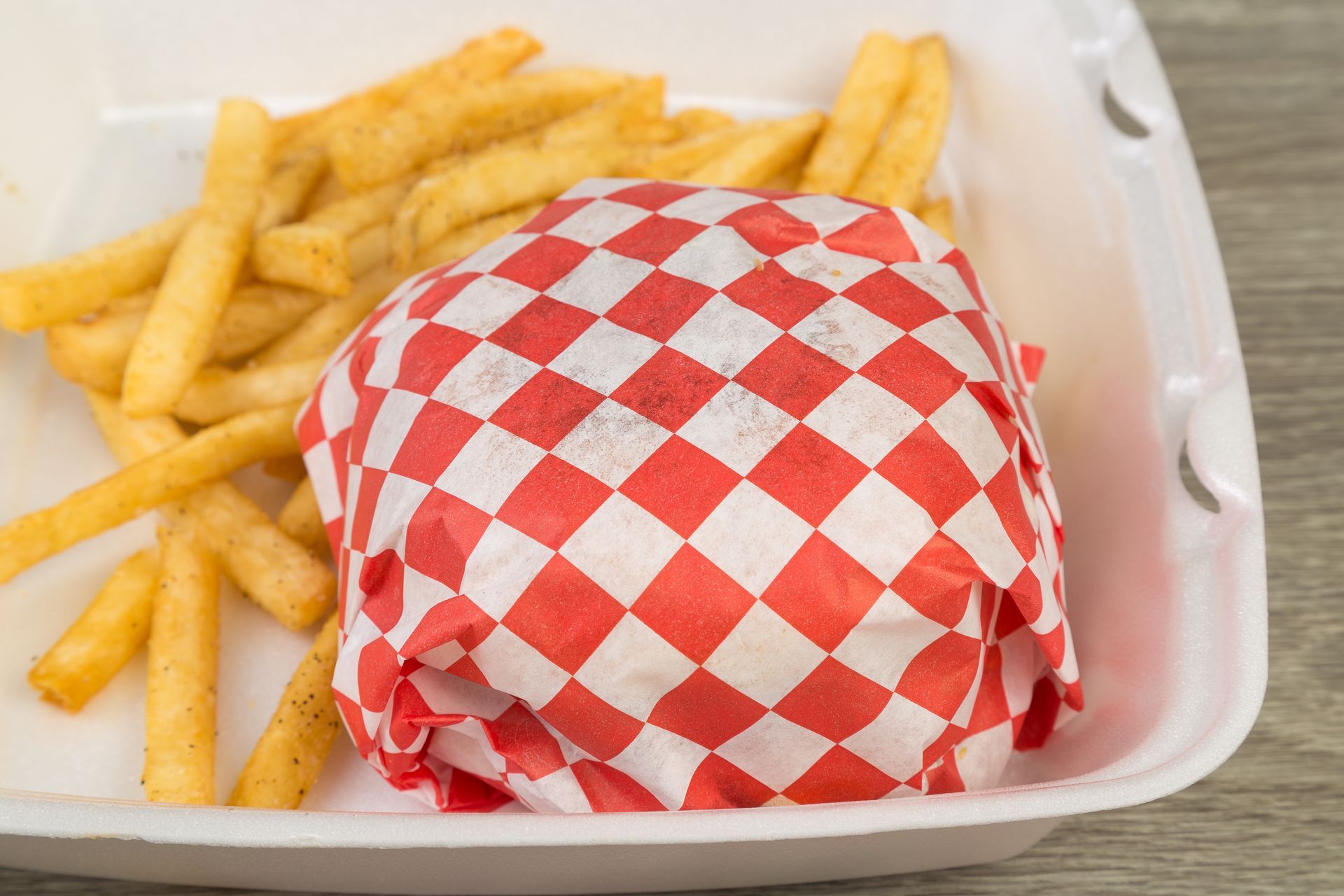 A hamburger wrapped in red and white checkered paper next to french fries in a styrofoam container.