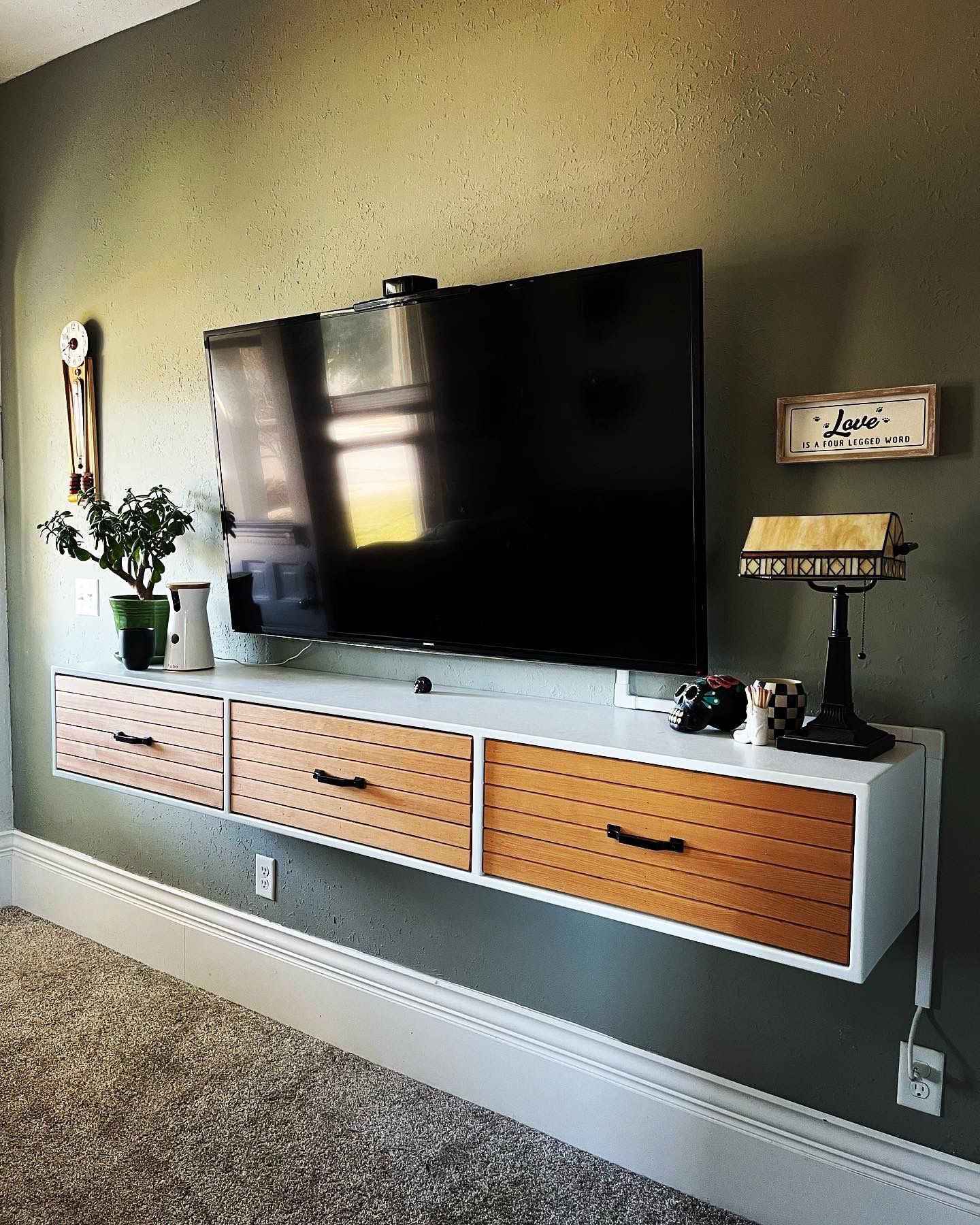 A wall-mounted white entertainment unit with three slatted wood drawers beneath a flat-screen TV on a gray-painted wall.