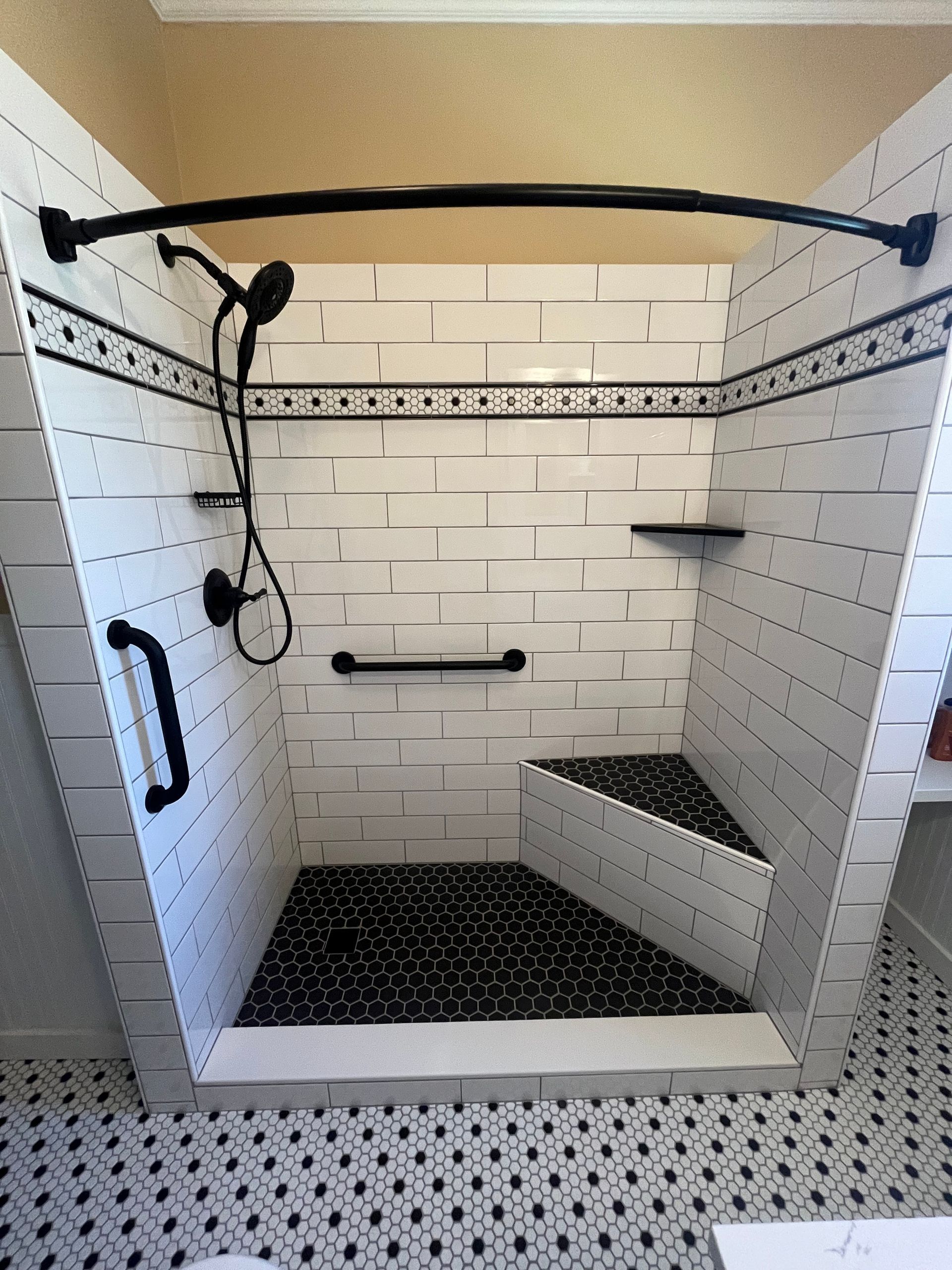 Walk-in shower with white subway tiles, a black pebble floor, corner bench, grab bars, and a black curved shower rod.