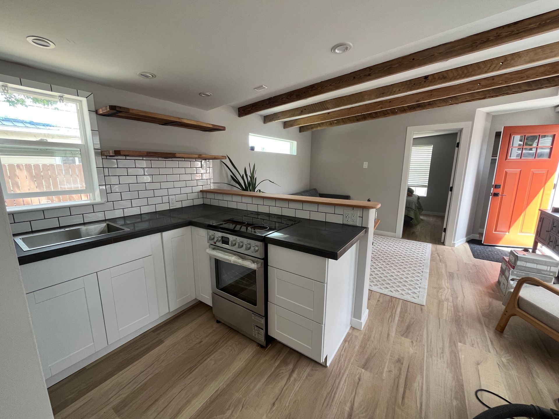 A modern kitchen with white cabinets, black countertops, wood open shelving, exposed ceiling beams, and an orange door.