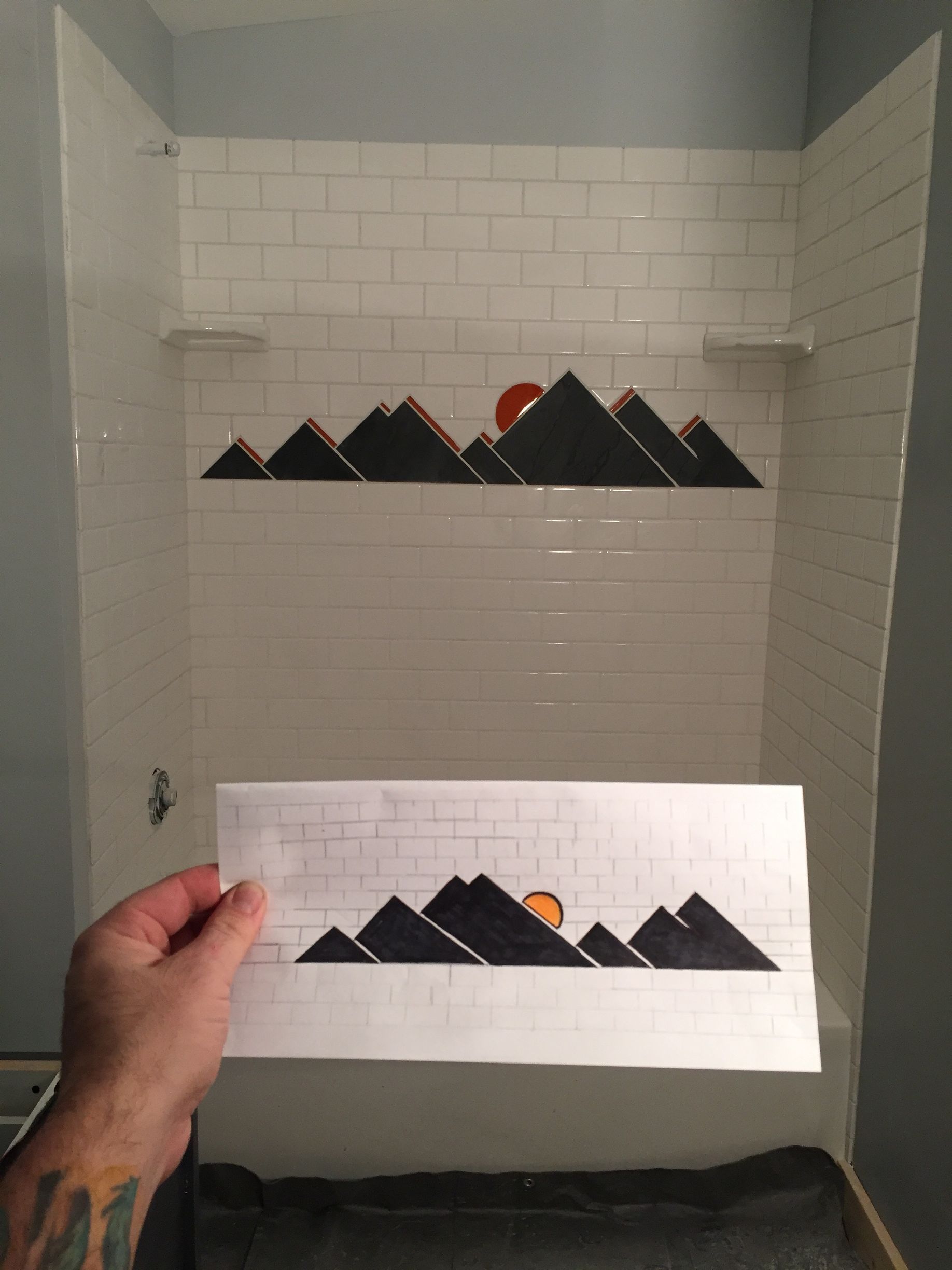 A hand holds a paper sketch of a mountain range with an orange sun in front of a tiled shower wall featuring the design.
