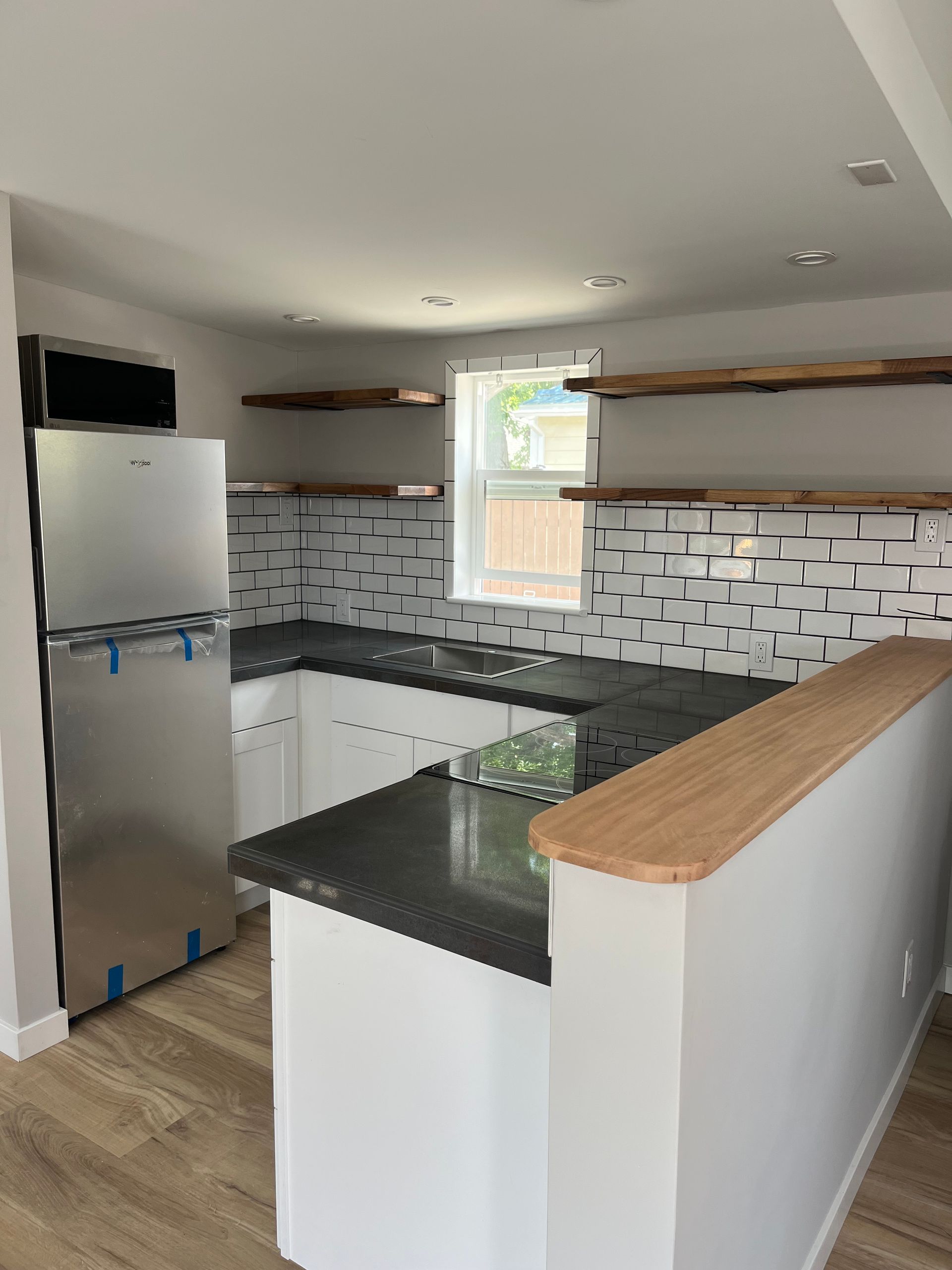 A modern kitchen featuring white cabinets, dark countertops, a stainless steel refrigerator, and wood shelving.