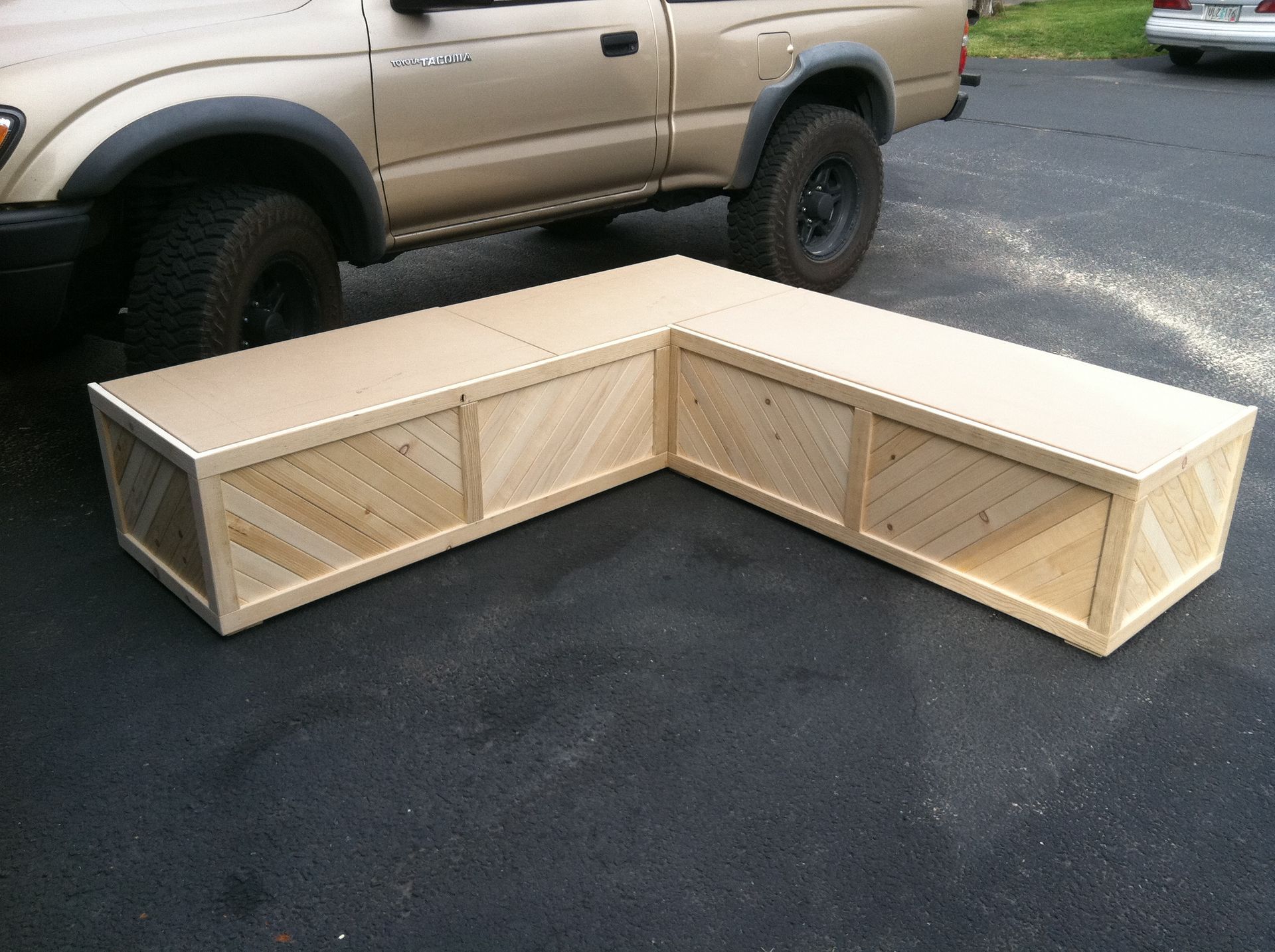 An L-shaped wooden bench with diagonal slat paneling, sitting on an asphalt driveway next to a tan pickup truck.