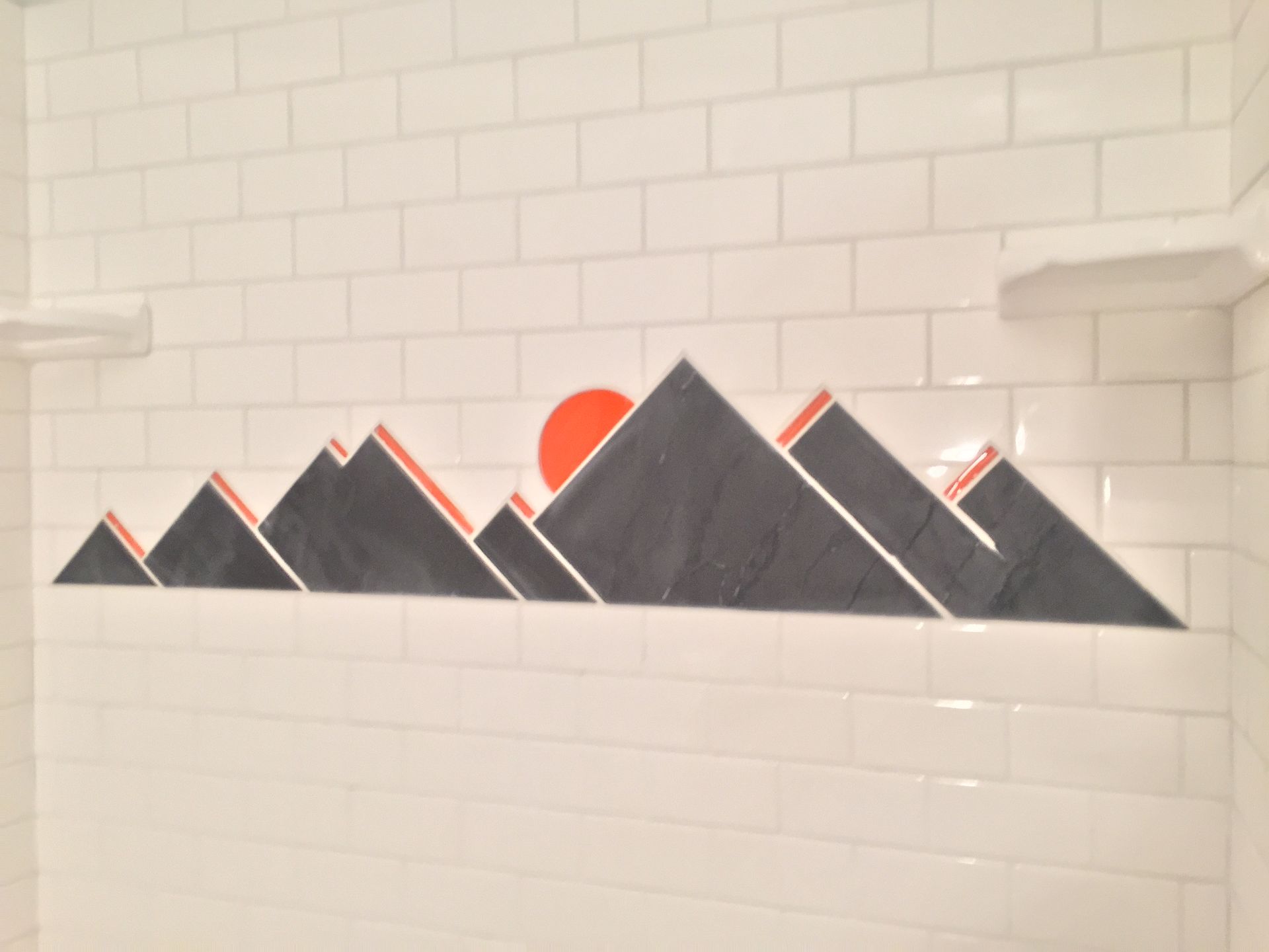 A decorative wall mural of dark gray mountains with orange sun and accents, set against a white subway tile background.