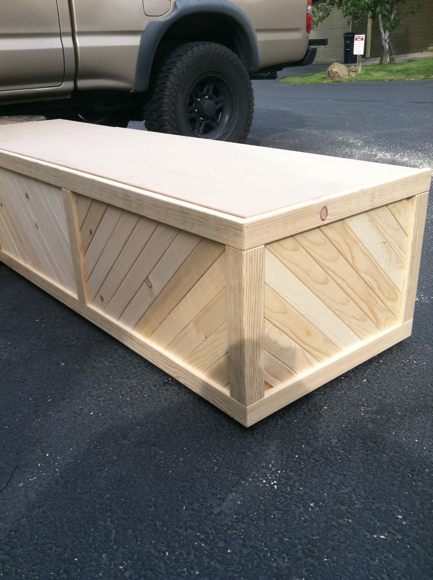 A custom wooden box with a diagonal plank design, sitting on asphalt next to a parked truck.