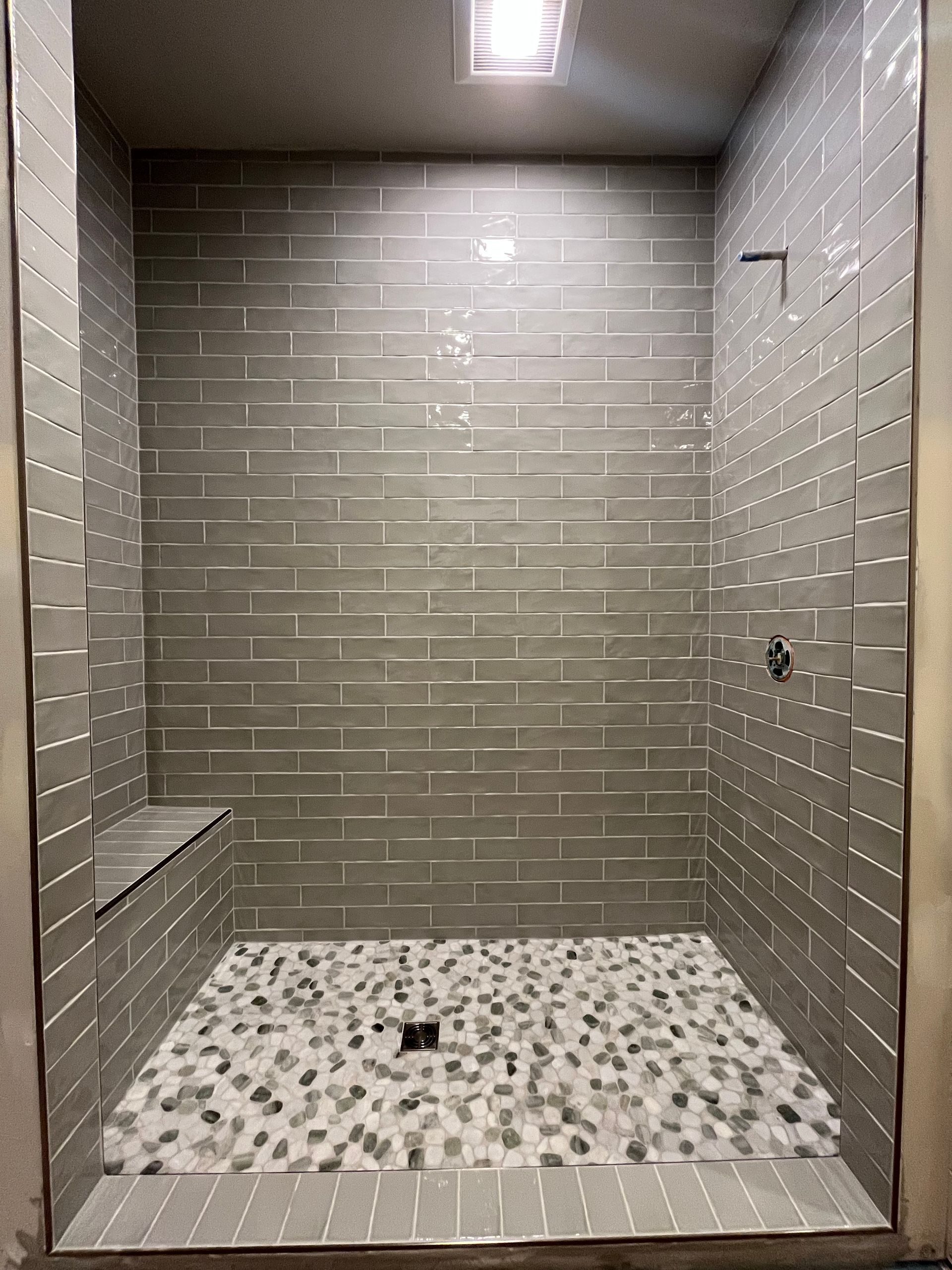 A walk-in shower with grey subway-tiled walls, a built-in corner seat, and a light-colored pebble floor.