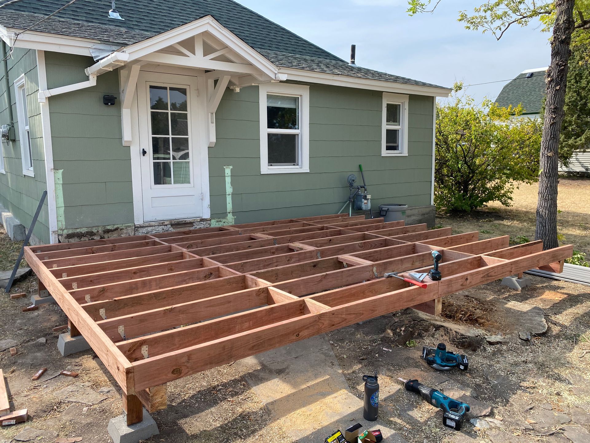 A wooden deck frame under construction in front of a green house, with power tools resting on the ground nearby.