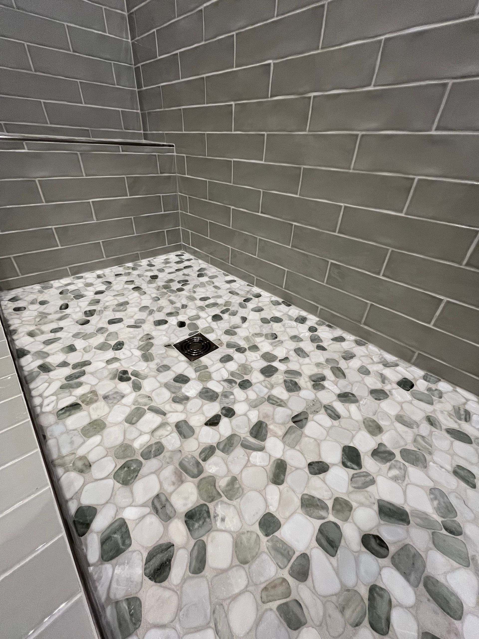 A shower floor with grey, white, and black river stone tiling, featuring a central square drain and dark grey wall bricks.