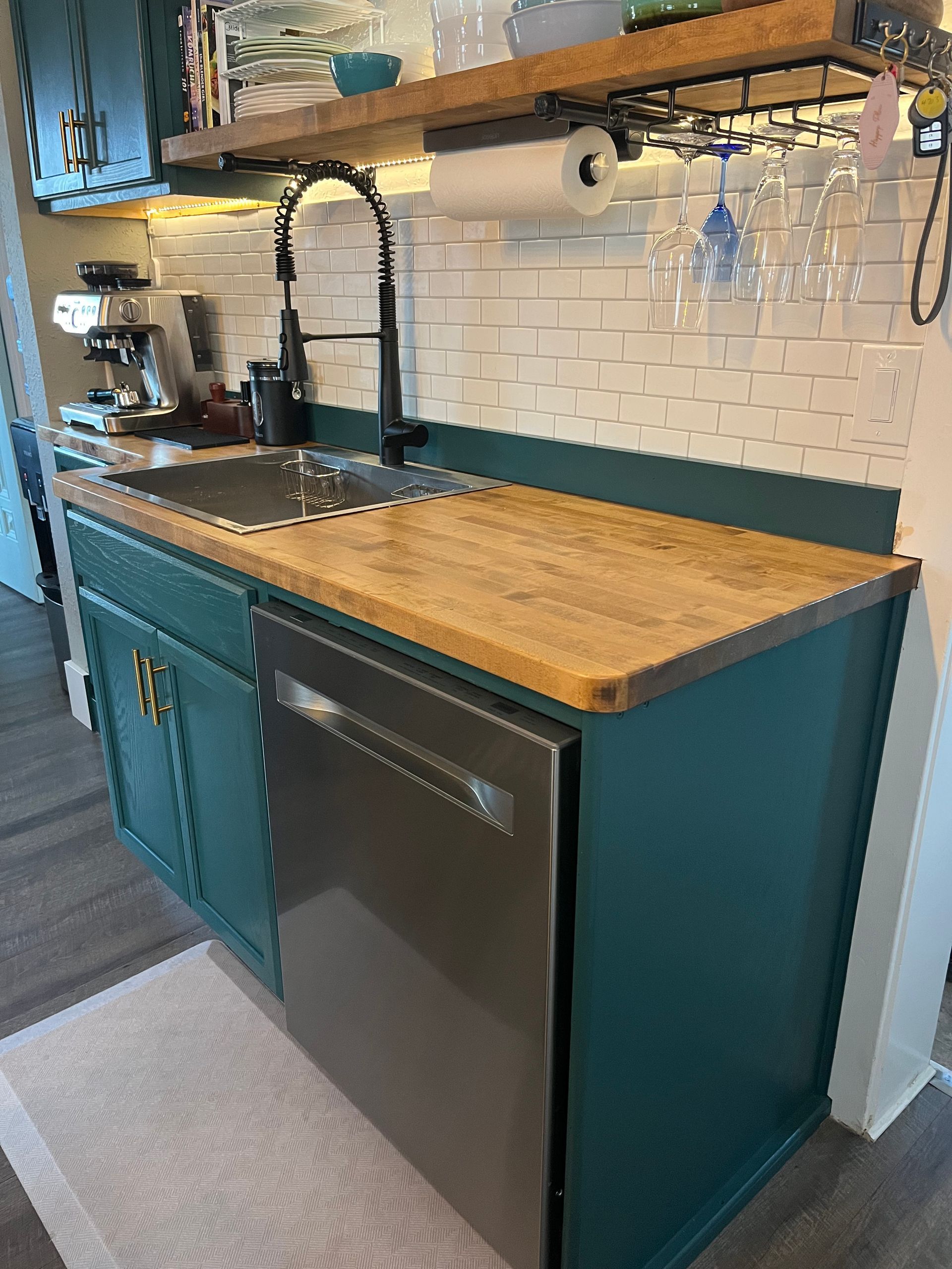 A dark teal kitchen cabinet base with a wooden countertop, a stainless steel dishwasher, and an open wooden shelf above.