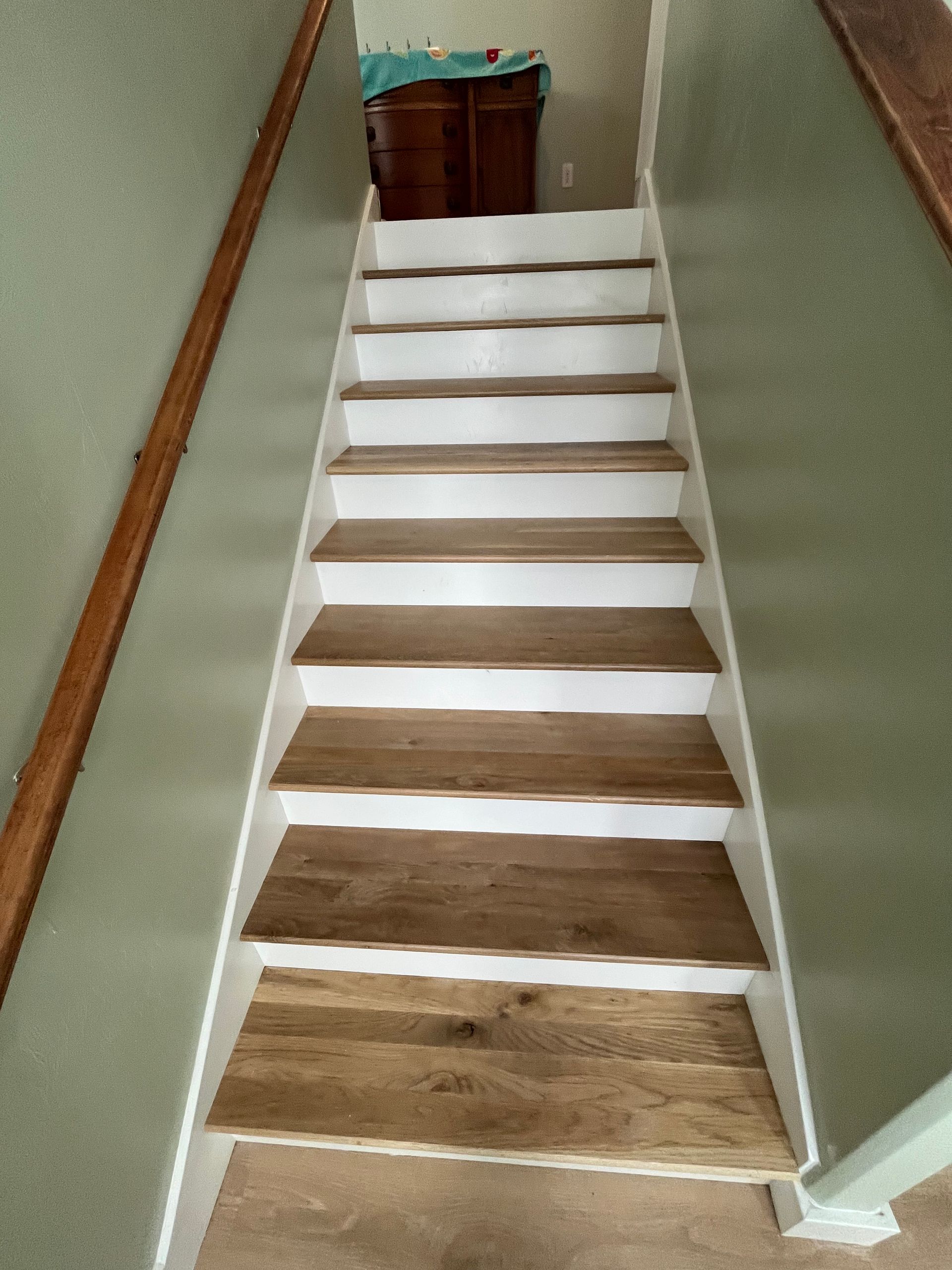 A straight wooden staircase with white risers and natural wood treads between two sage green walls.