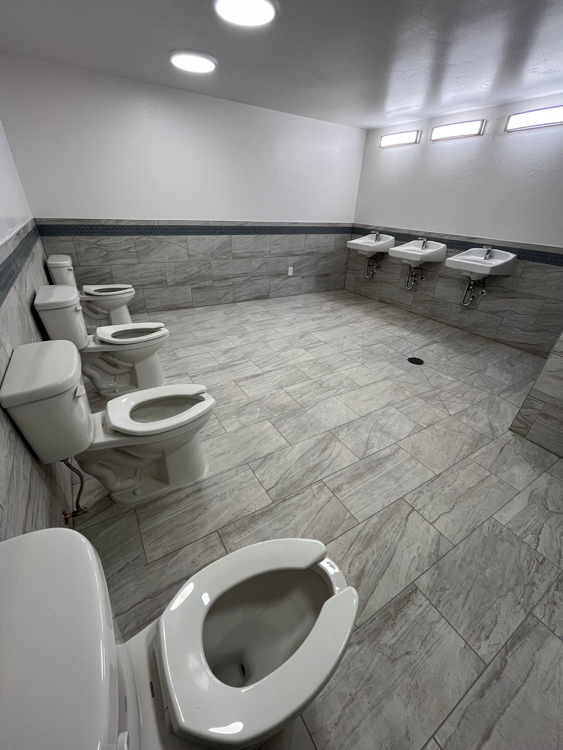 A communal restroom with multiple open toilets and sinks, featuring light grey stone-tiled walls and floors.
