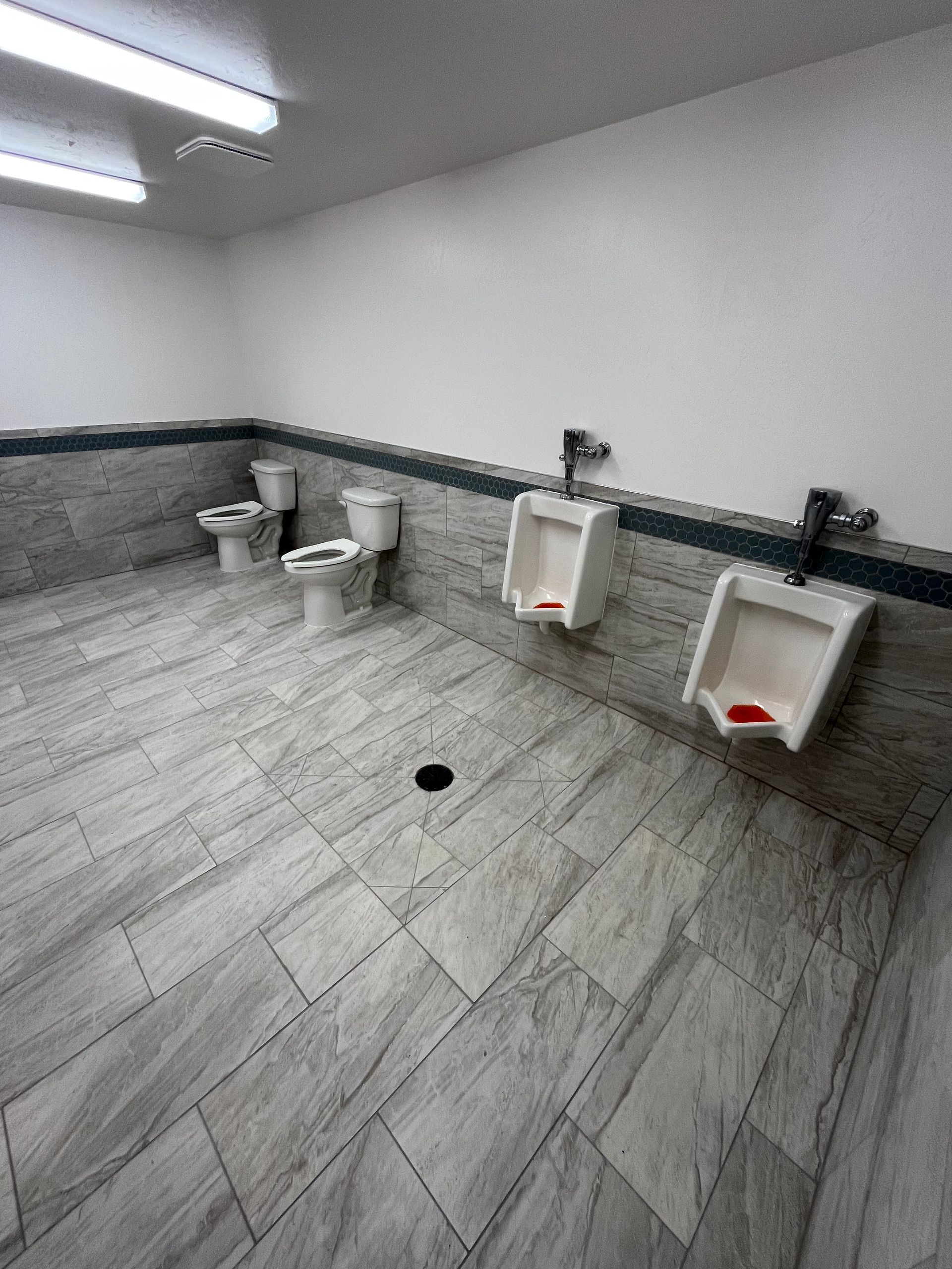 A modern restroom with two wall-mounted toilets and two urinals against a tiled wall with light gray stone-patterned floors.
