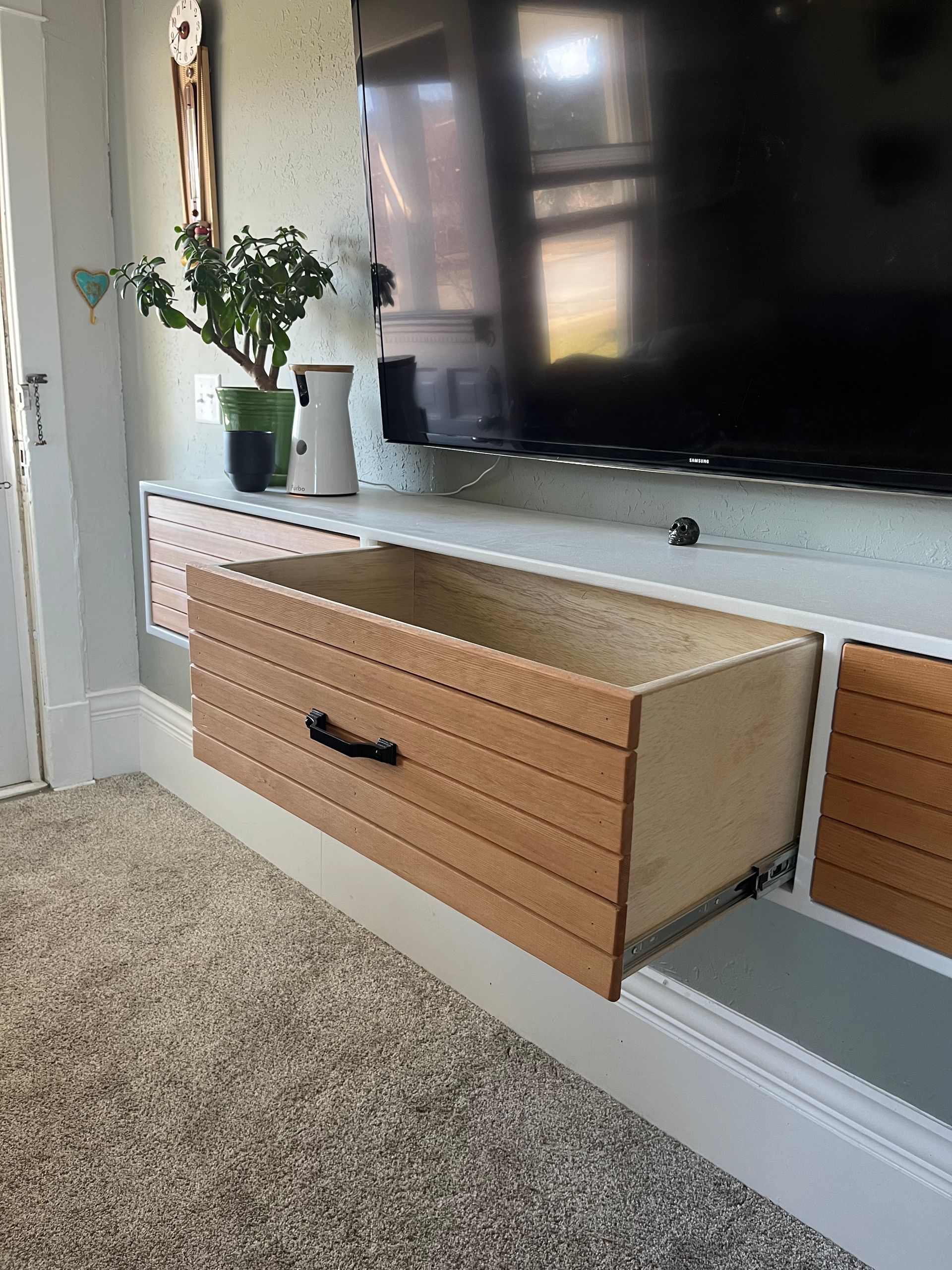 A floating shelf under a TV with a pull-out wooden drawer and a small green plant on top.