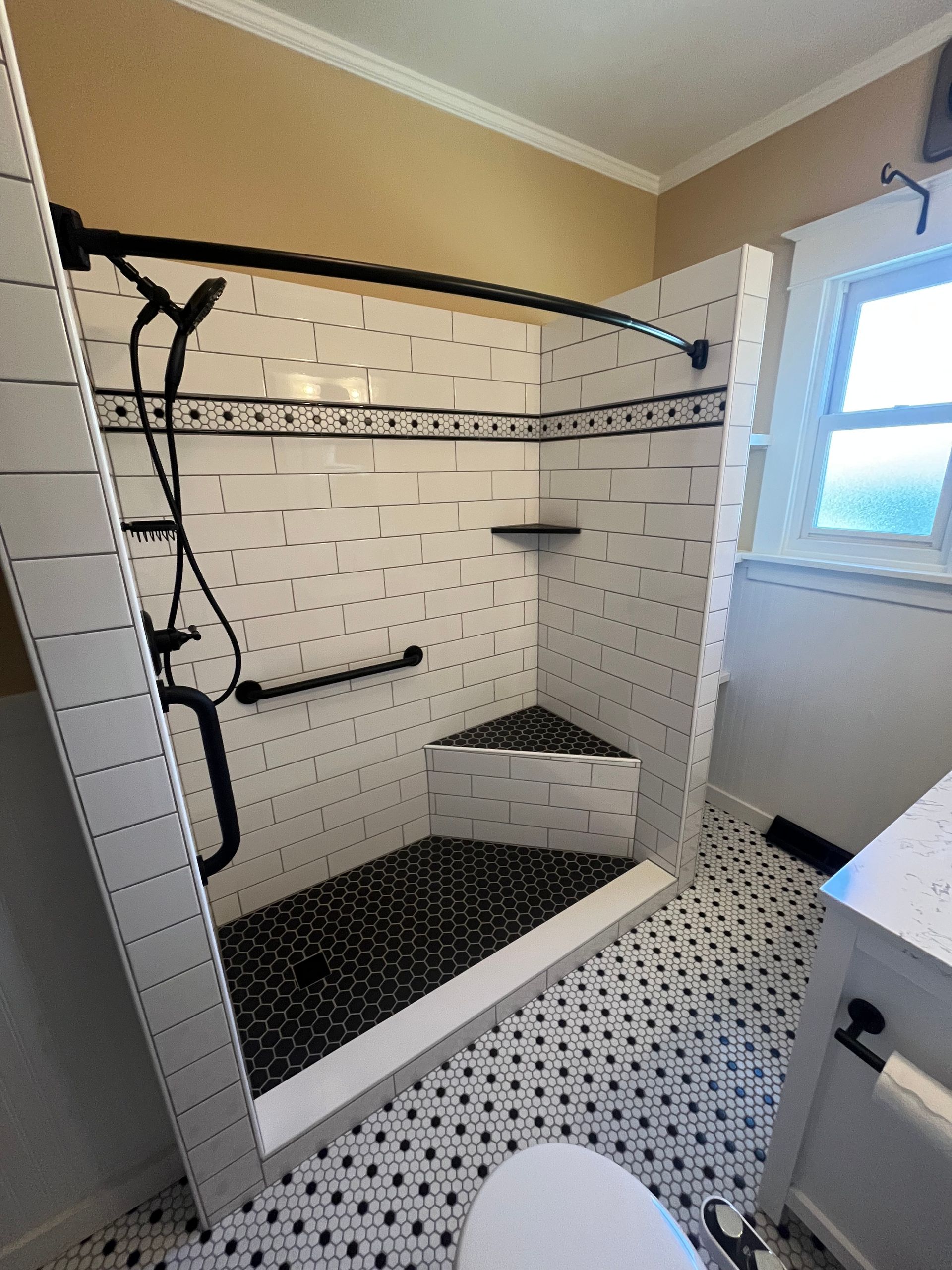 Walk-in shower with black-and-white tile, a built-in corner seat, a safety grab bar, and a handheld showerhead.
