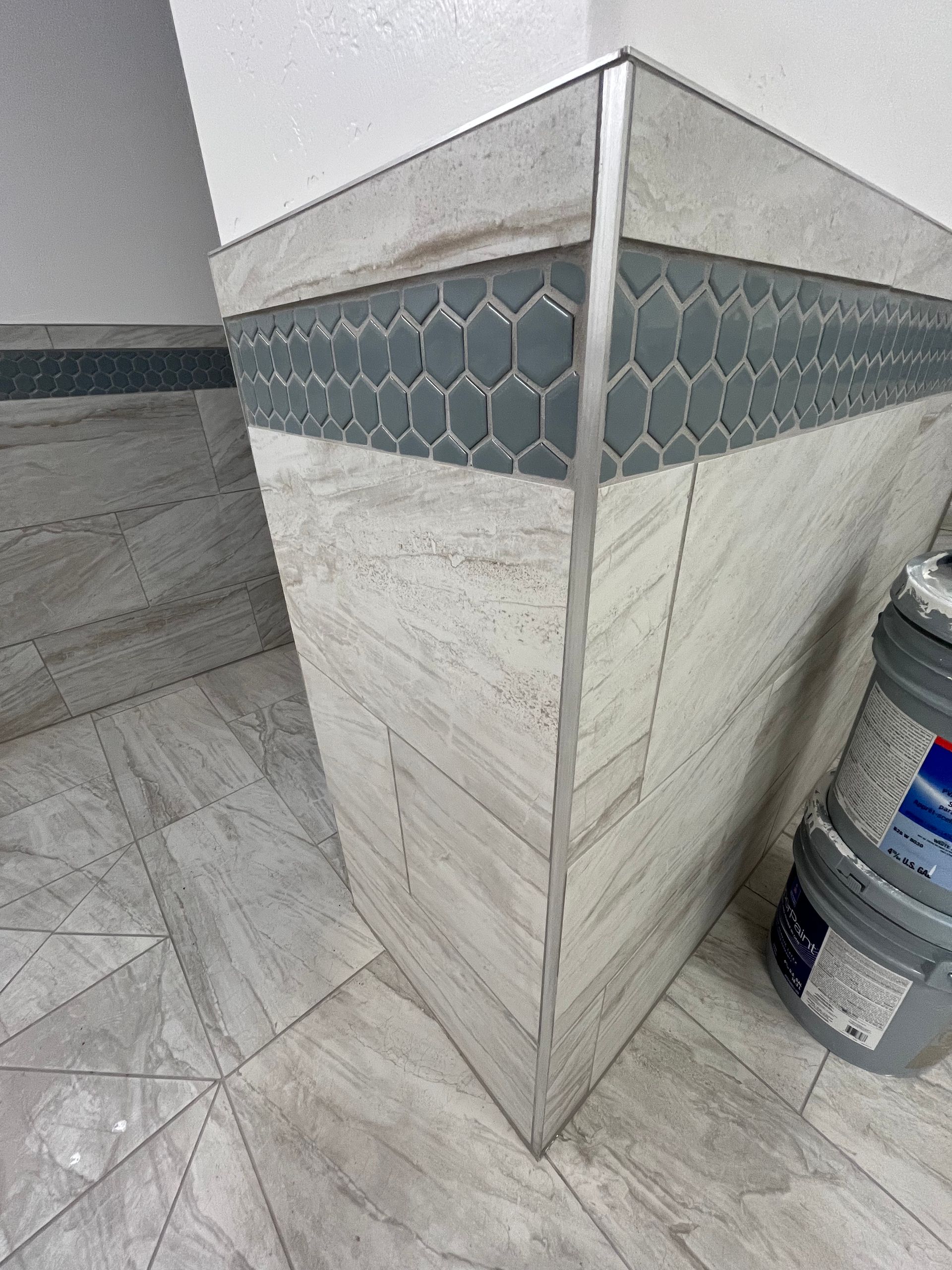 A corner of a tiled wall featuring gray hexagon mosaic accents, wood-look beige tiles, and a metal corner trim piece.