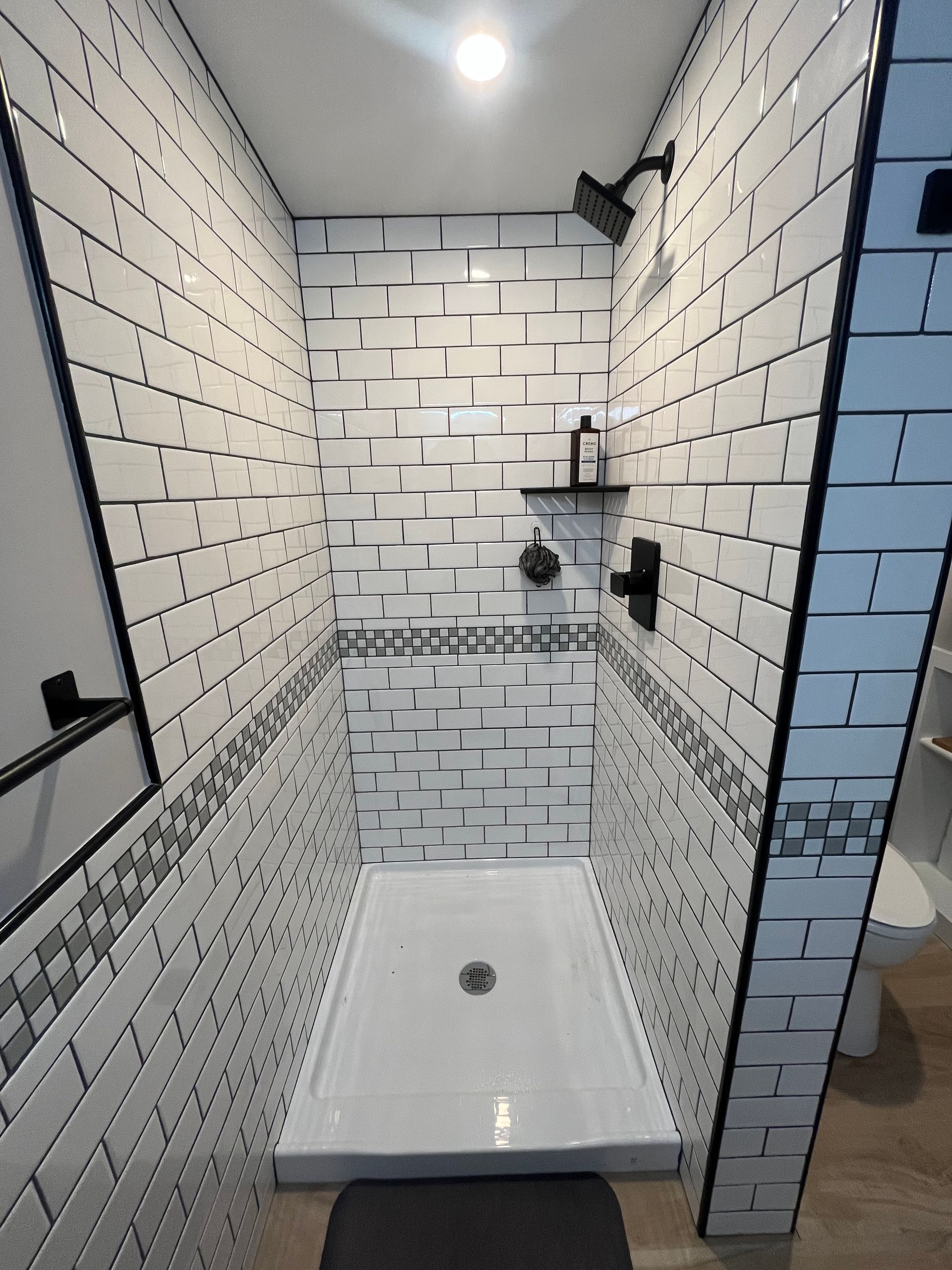 A walk-in shower with white brick-style tiles, dark grout, a black shower head, and a triangular corner shelf.