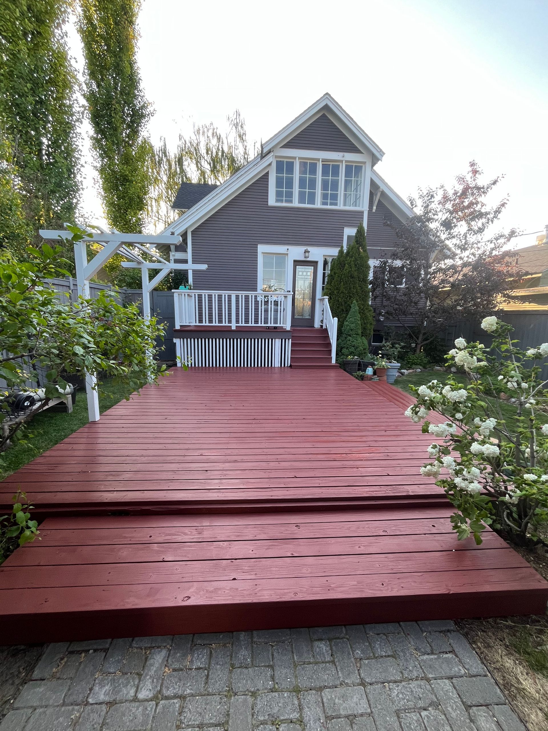 A two-story gray house with a large, two-tiered red wooden deck, white railings, and a stone walkway in a garden setting.