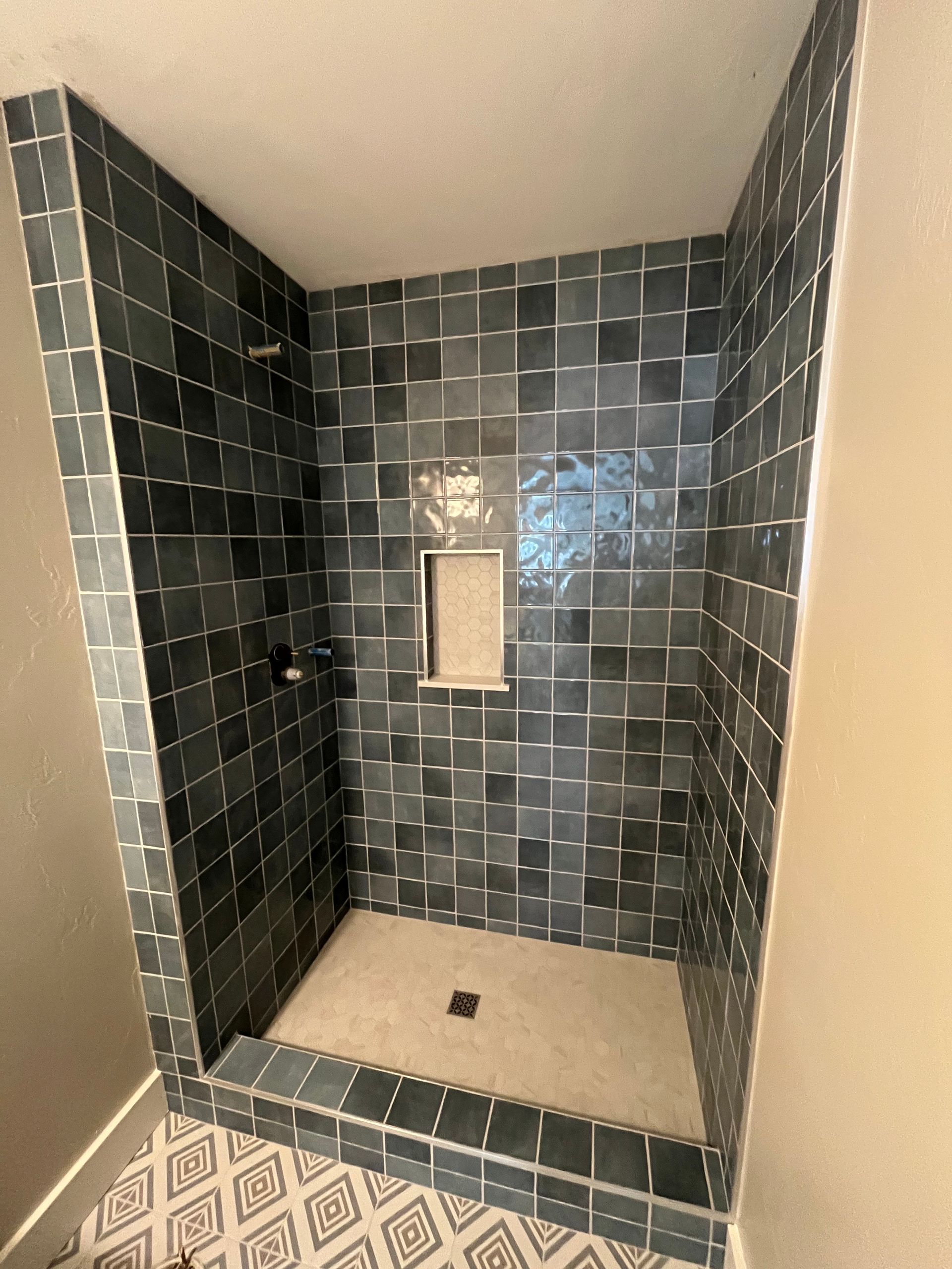 A shower stall with dark, grid-patterned tiles, a recessed wall niche, a light-colored floor, and a patterned rug in front.