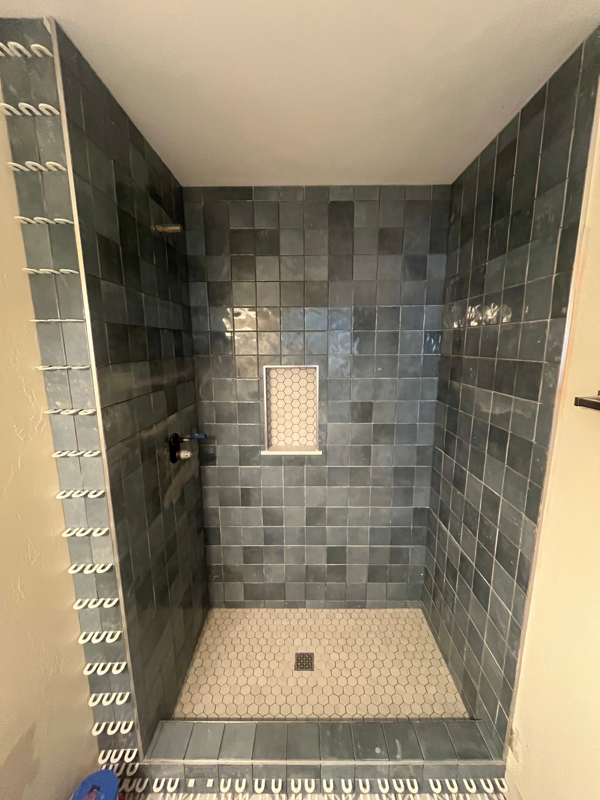 A shower stall featuring dark gray, rectangular wall tiles, a light-colored stone floor, and a small built-in wall niche.