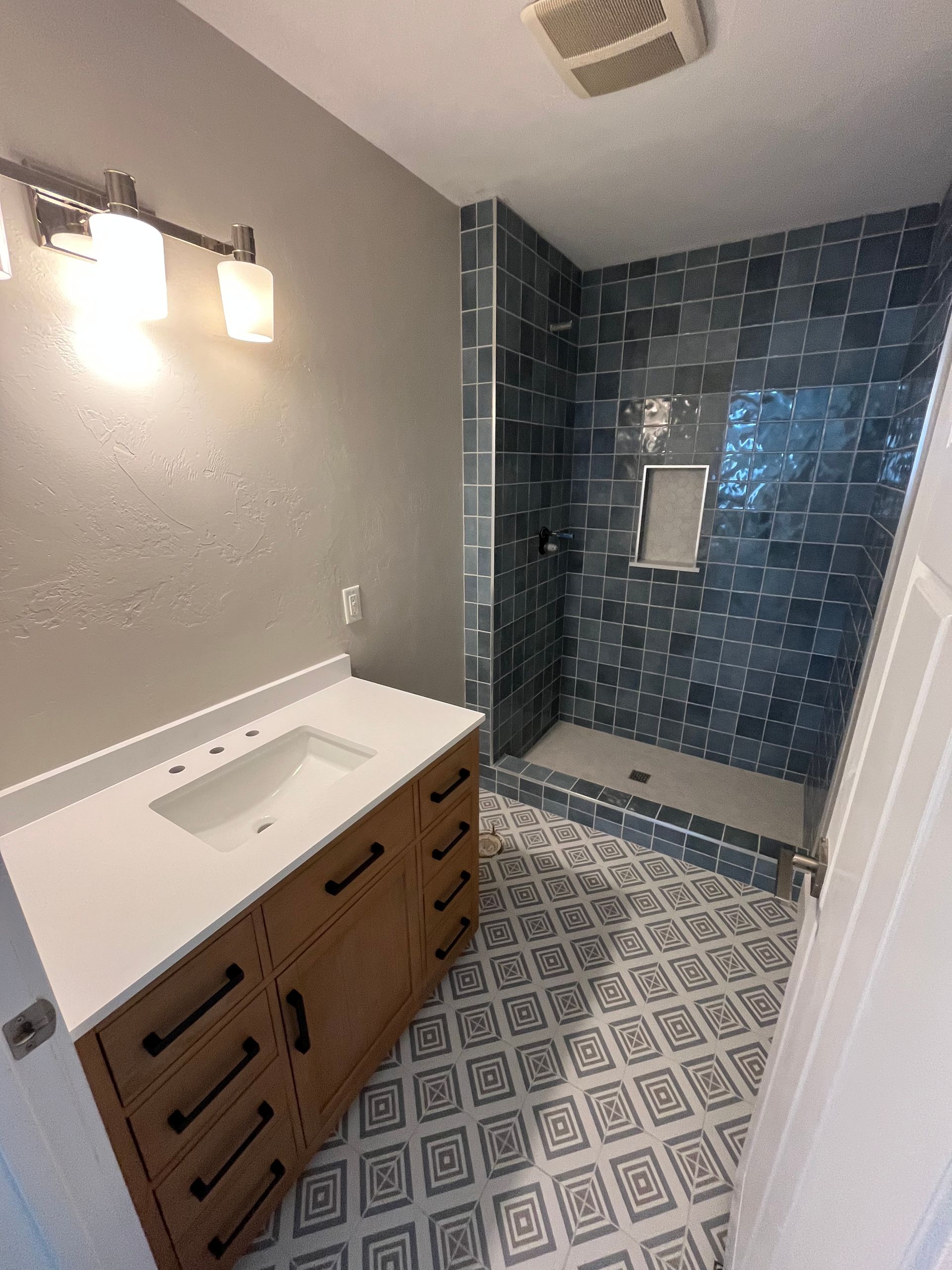 Bathroom with a wooden vanity, white countertop, patterned tile floor, and a walk-in shower with dark blue square tiles.