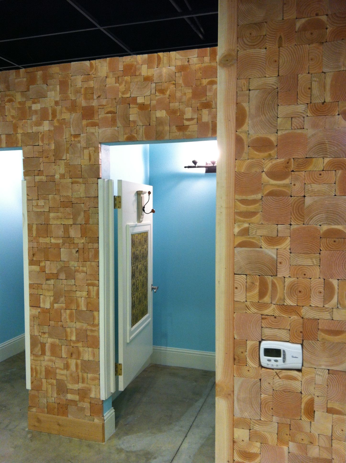 A wall covered in small wooden blocks features a doorway into a blue-walled restroom with a white door and thermostat.