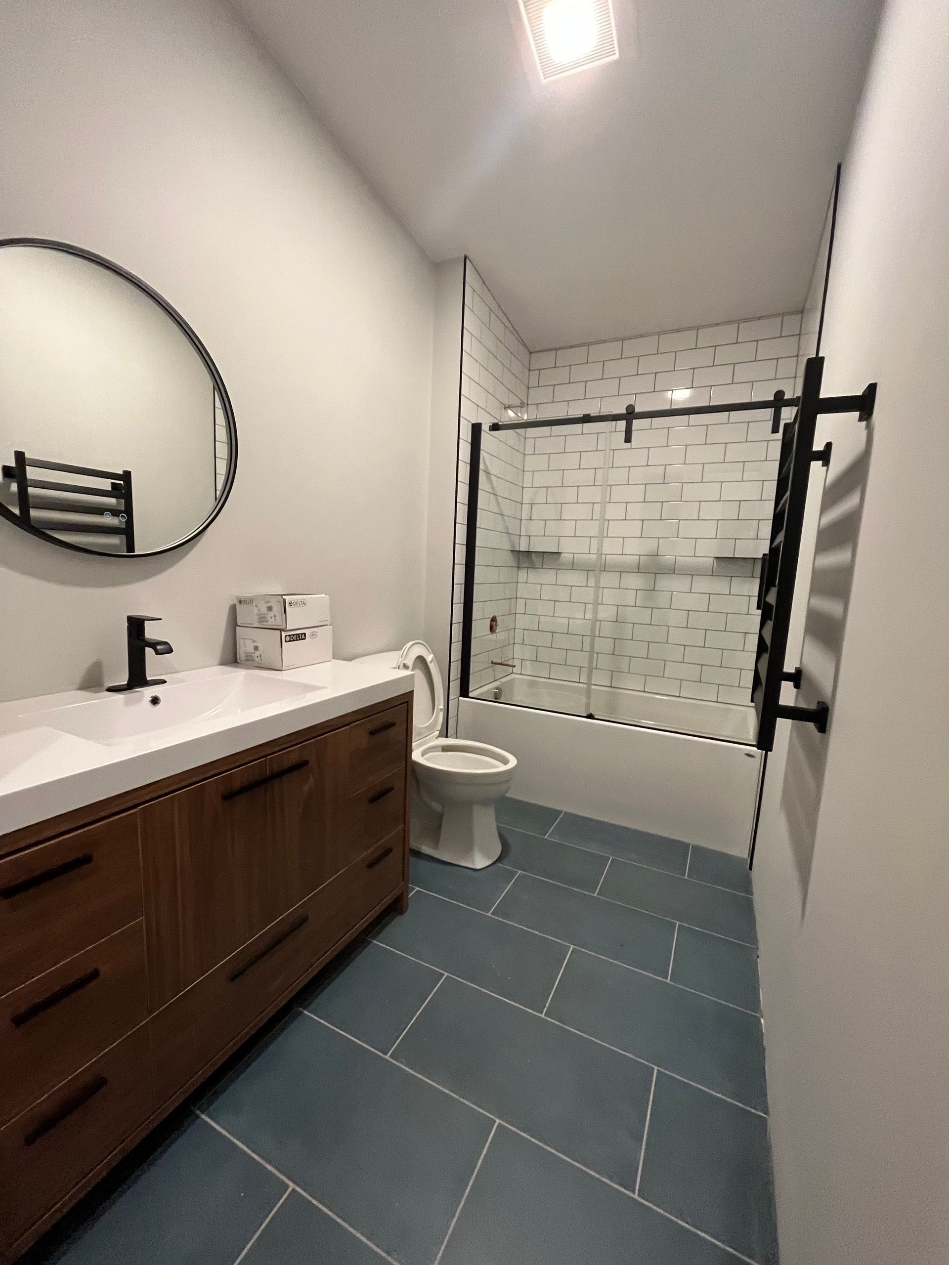 Modern bathroom with a wooden vanity, a round mirror, blue tiled flooring, and a glass-enclosed shower with white tiles.